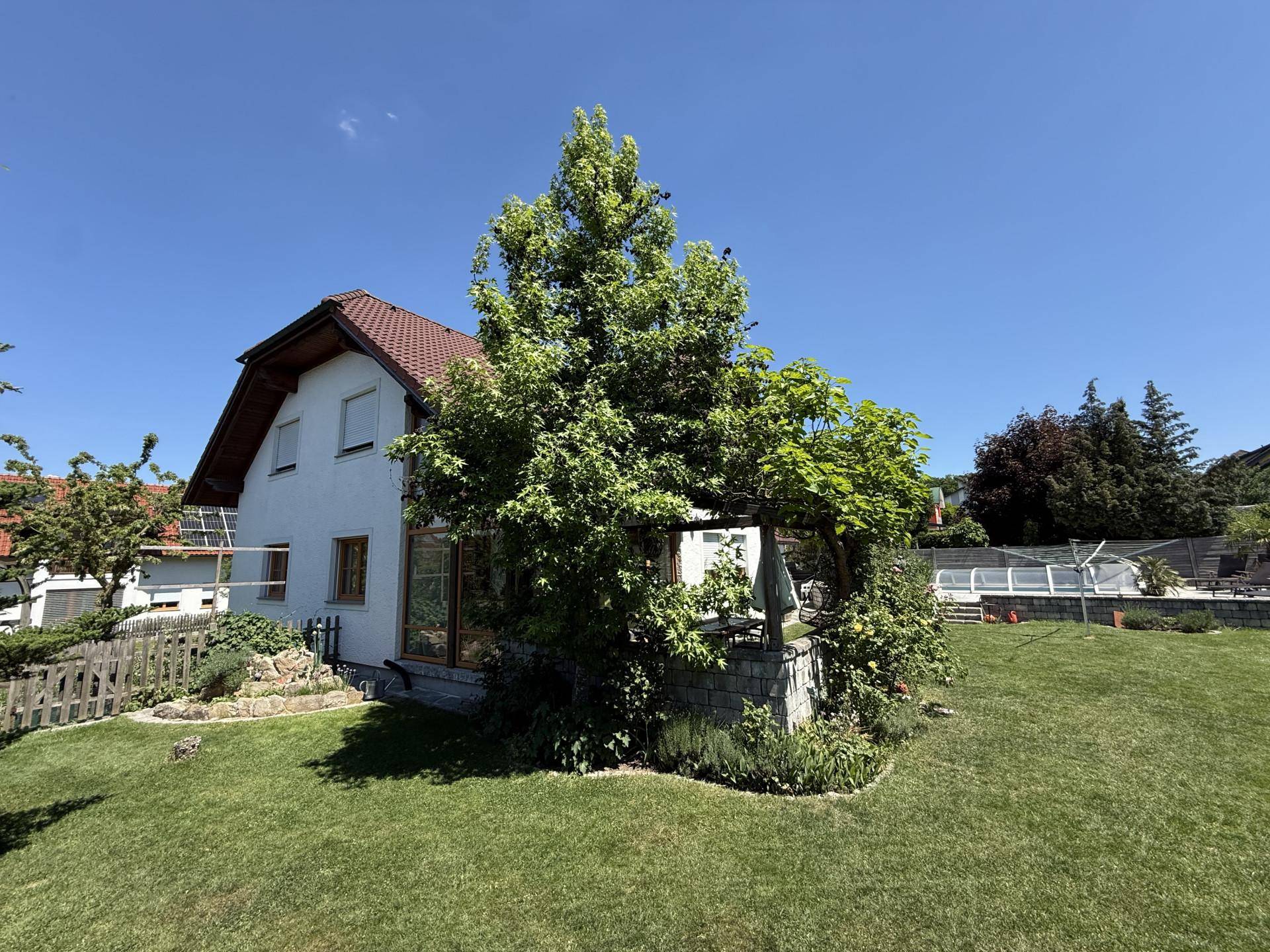 Seitenansicht des Wohnhauses mit großem Baum, gepflegtem Garten und Poolbereich im Hintergrund.