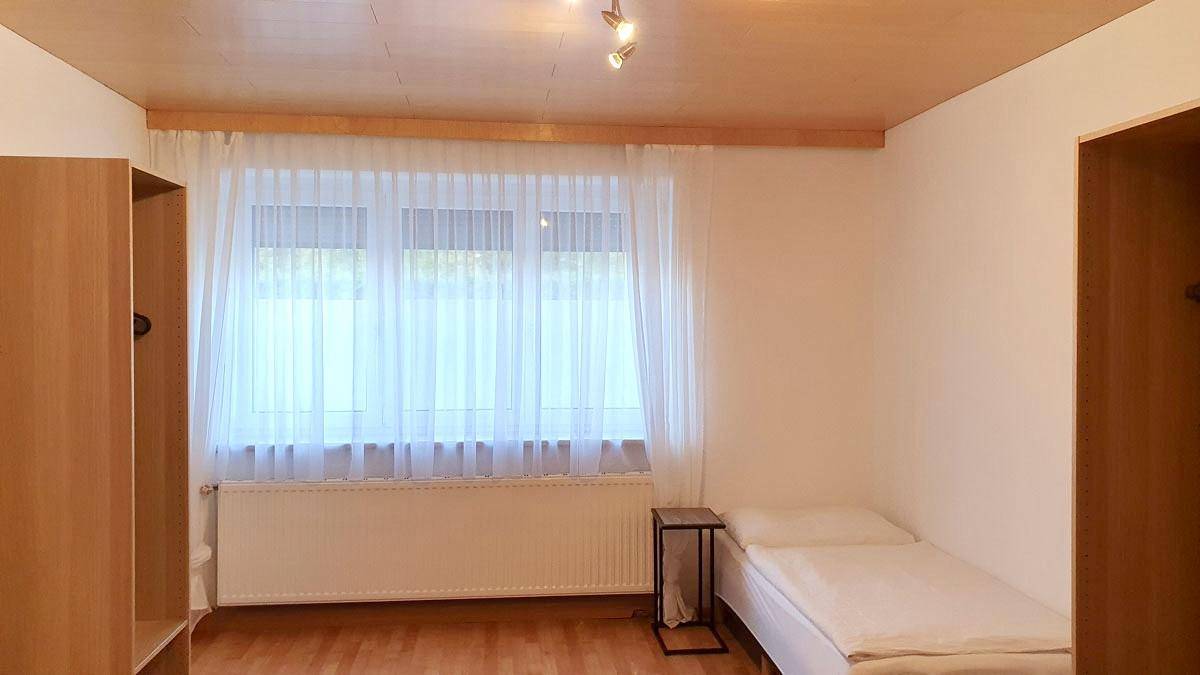 Einfaches Schlafzimmer mit Einzelbett, Holzfußboden und großem Fenster für viel Tageslicht.