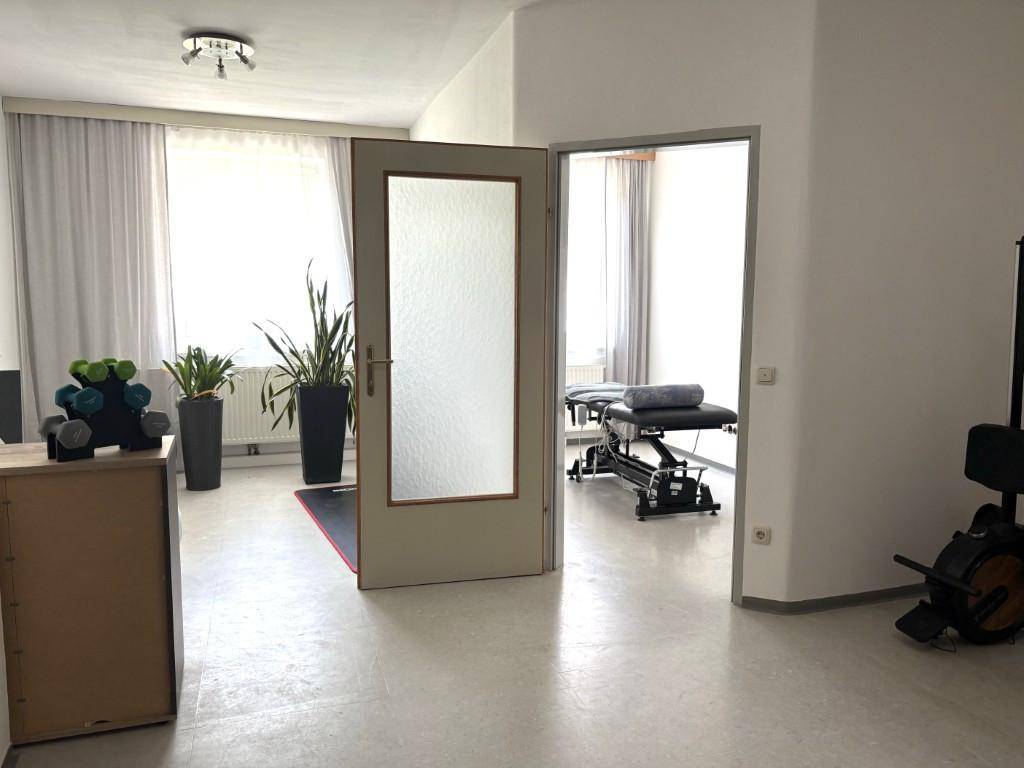 Zwei helle Räume, einer mit Fitnessgeräten und Pflanzen, der andere mit einer Massageliege.