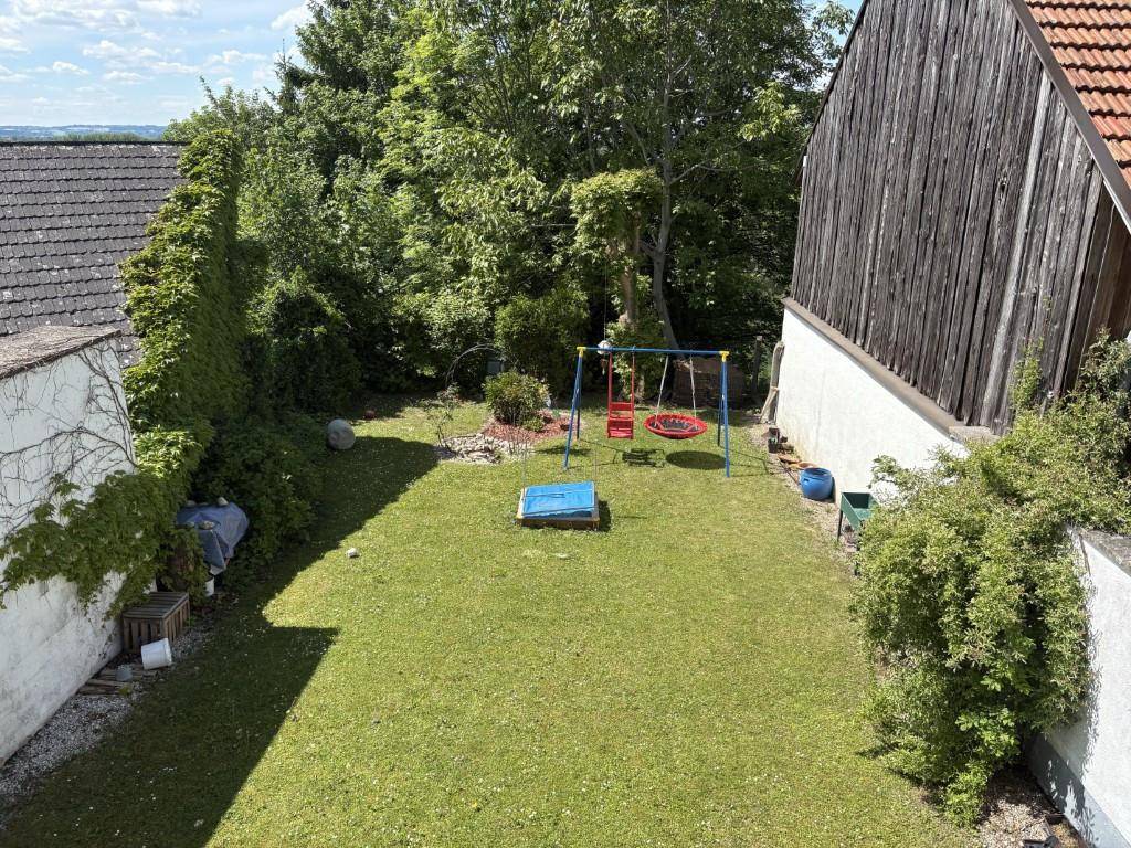 Grüner Gartenbereich mit Schaukel und Rutsche, umgeben von Bäumen und einer Mauer.