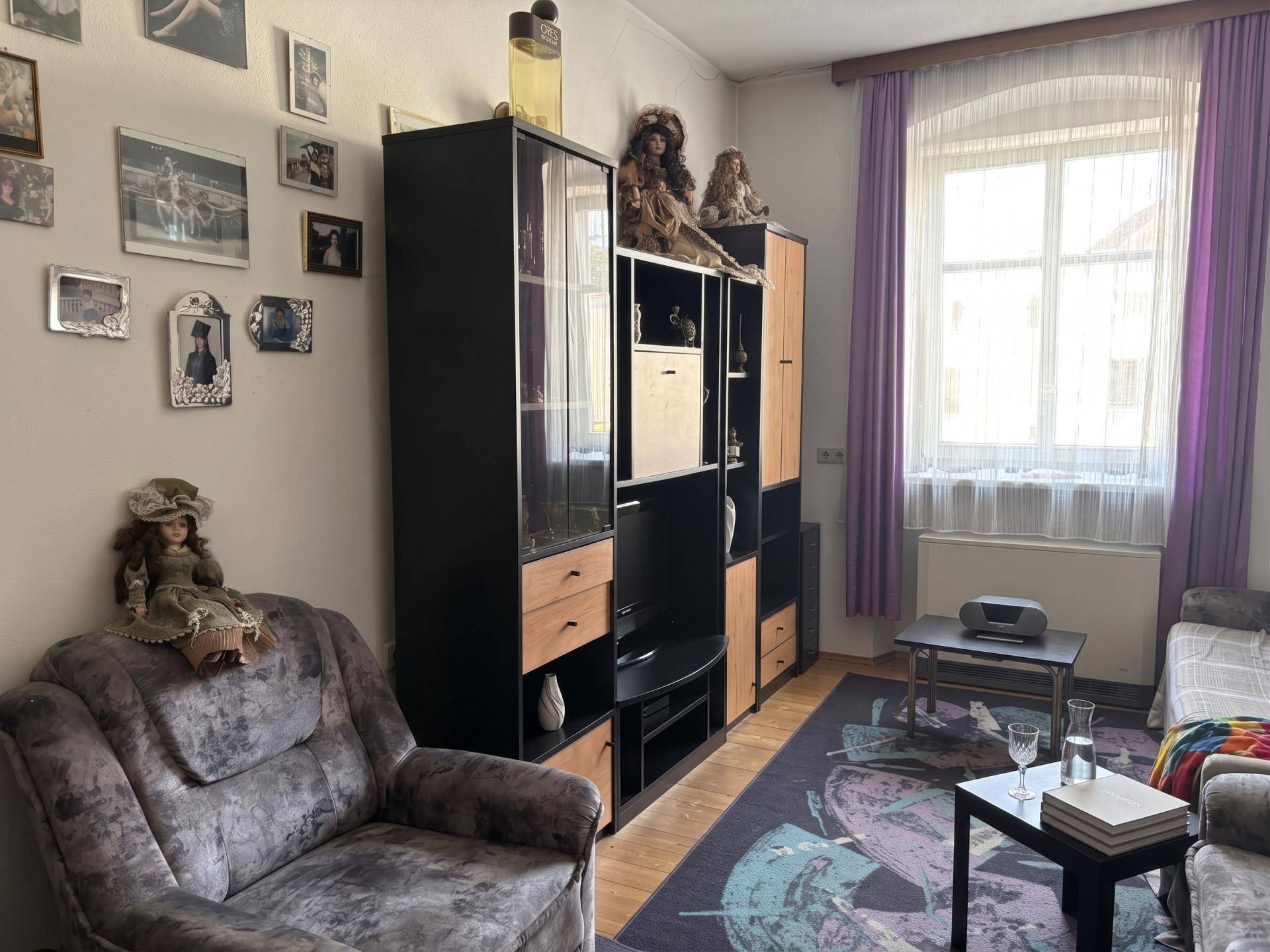 Gemütliches Wohnzimmer mit großer Schrankwand, Sessel und einem Fenster mit lila Vorhängen.