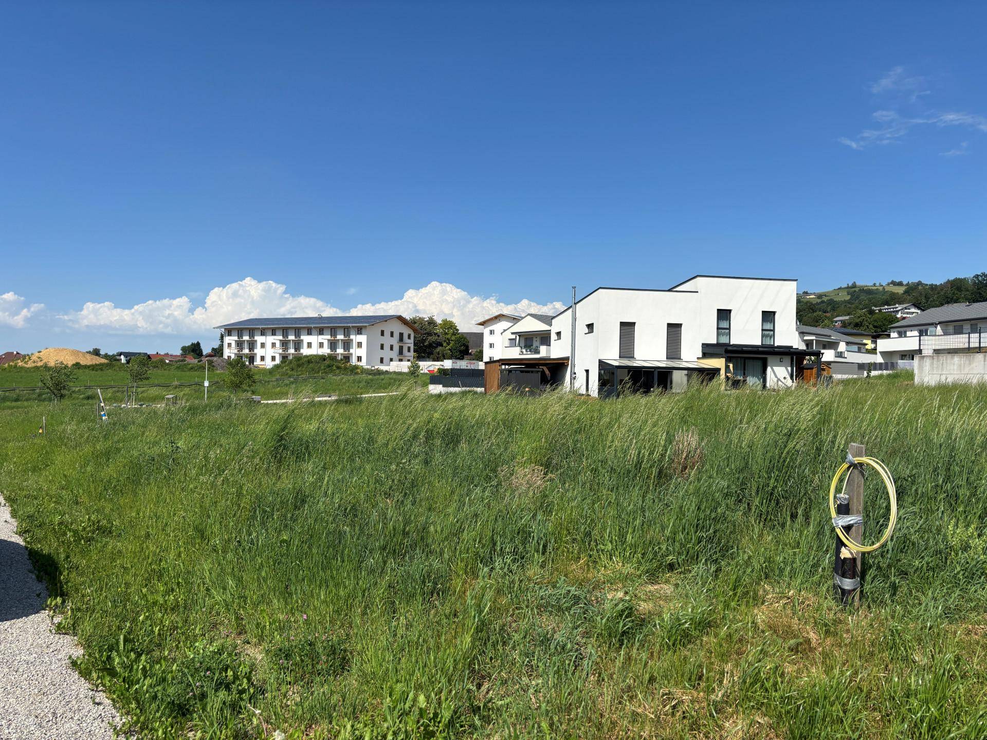 Blick über eine grüne Wiese auf moderne weiße Gebäude und eine Wohnsiedlung unter blauem Himmel.