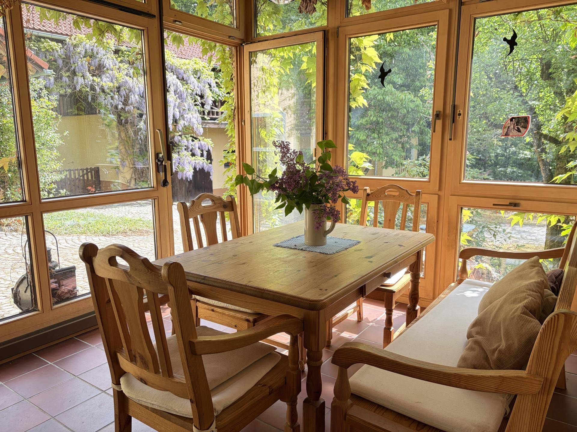 Heller Wintergarten mit Holztisch und Stühlen, umgeben von großen Fenstern mit Blick ins Grüne.