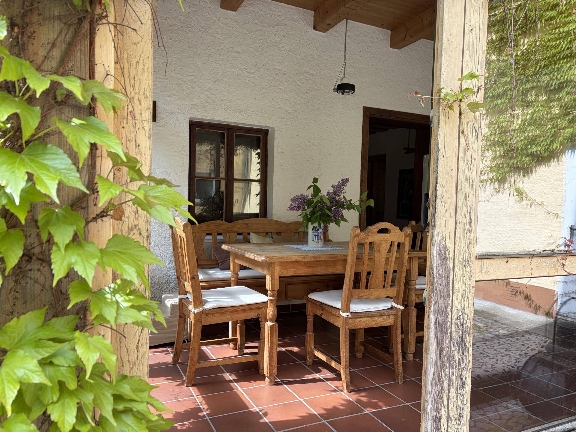 Überdachte Terrasse mit Holztisch und Stühlen, umgeben von Kletterpflanzen, ideal für Mahlzeiten im Freien.