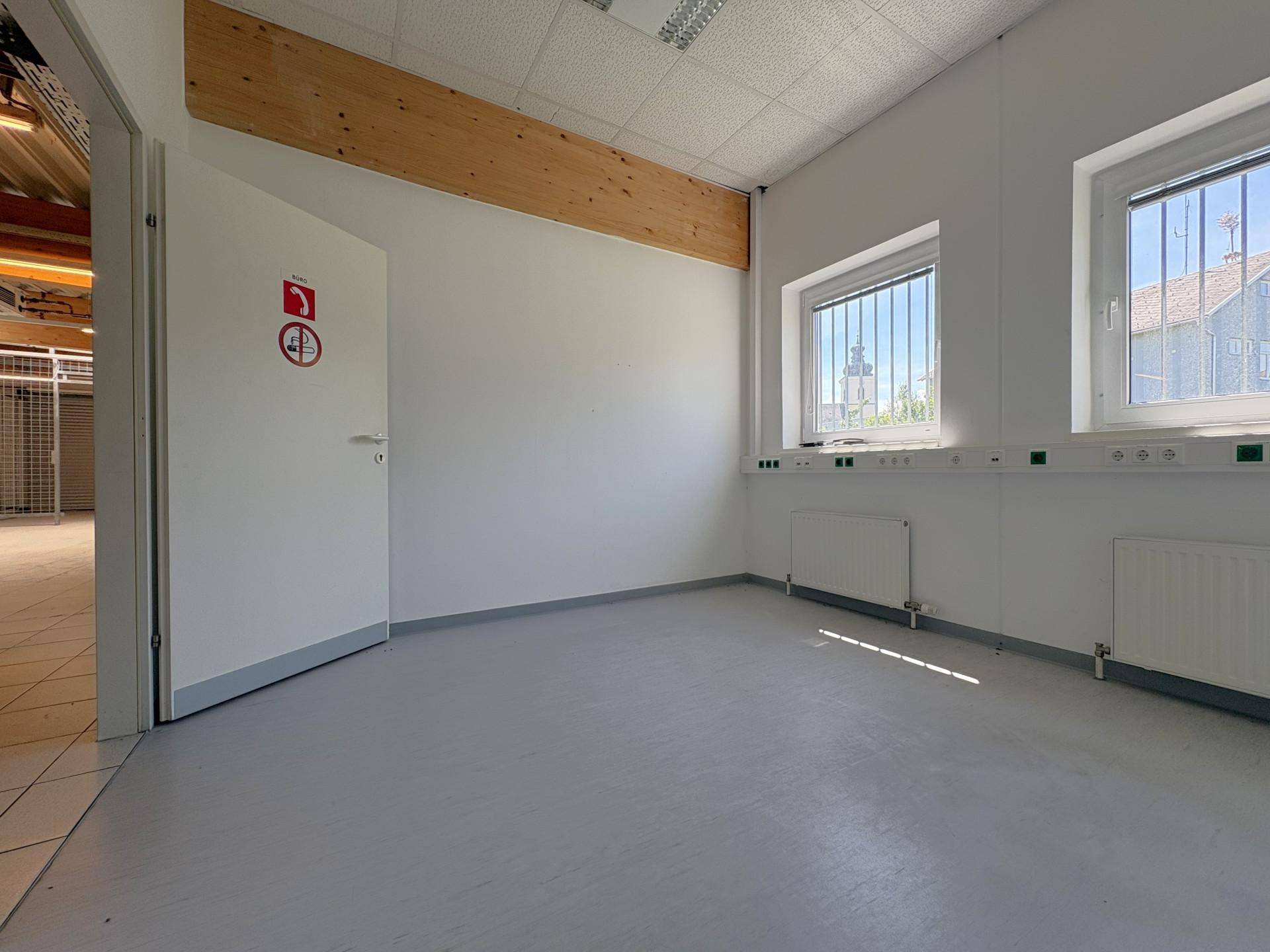 Kleiner Büroraum mit zwei Fenstern, Heizkörpern und hellem Vinylboden, einfach und funktional.
