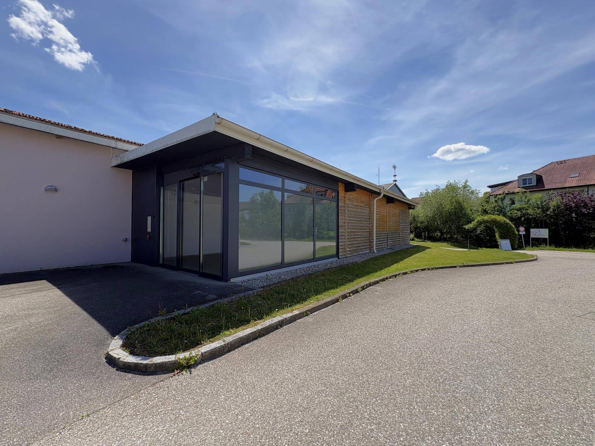Moderne Gebäudeecke mit großen Glasfronten und Holzverkleidung, umgeben von Grünflächen und Parkplatz.