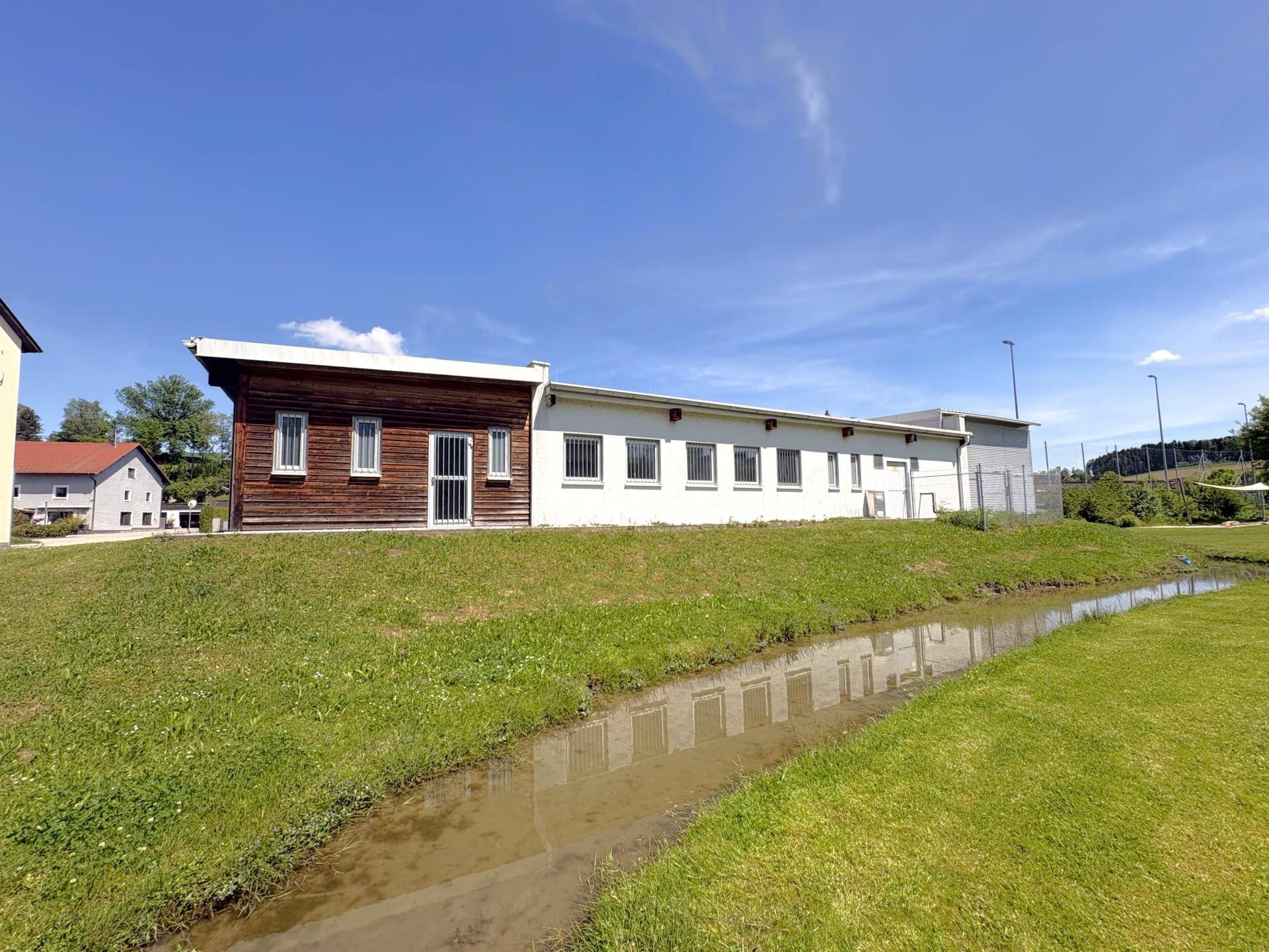 Gebäudeansicht mit Holzfassade und weiß verputztem Teil, umgeben von Grünflächen und einem kleinen Bach.