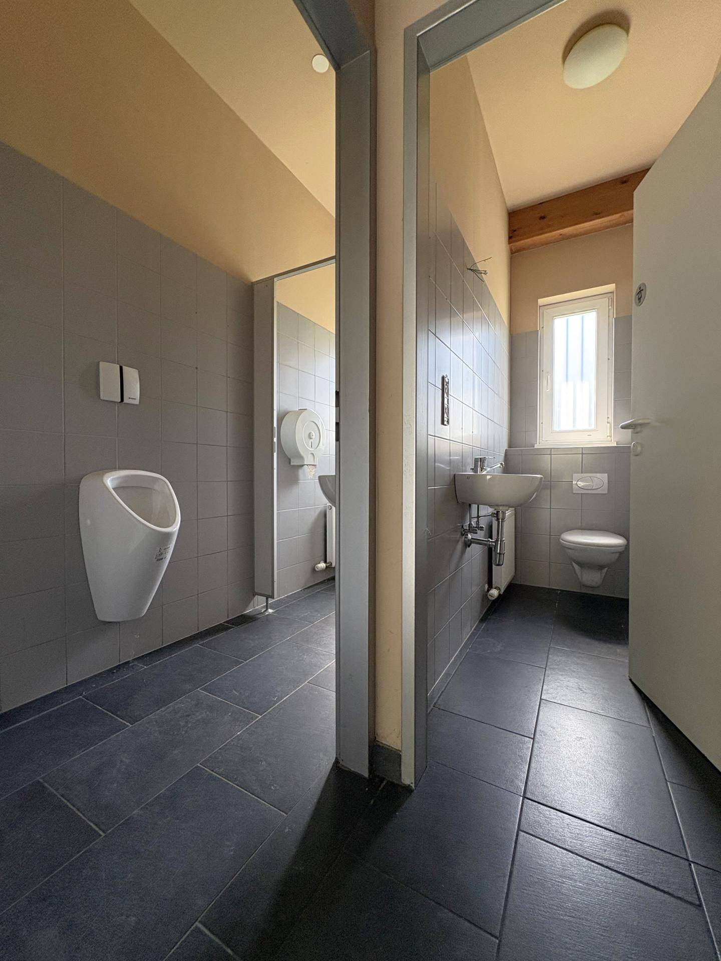 Öffentliche Toilettenanlage mit Urinal, Waschbecken und dunklen Fliesen, funktional und sauber.