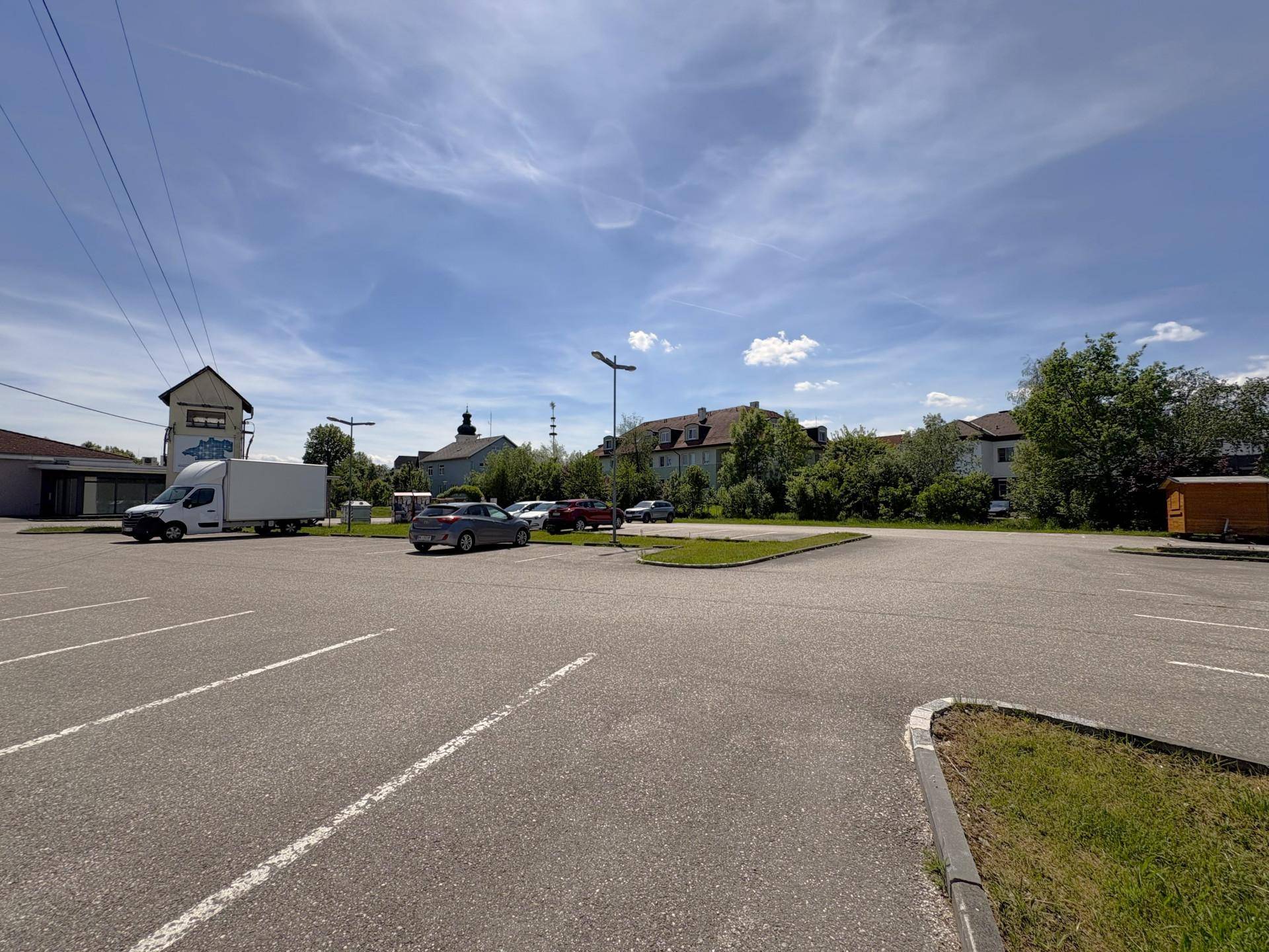 Weitläufiger Parkplatz mit Fahrzeugen und Gebäuden im Hintergrund, umgeben von Grünflächen.