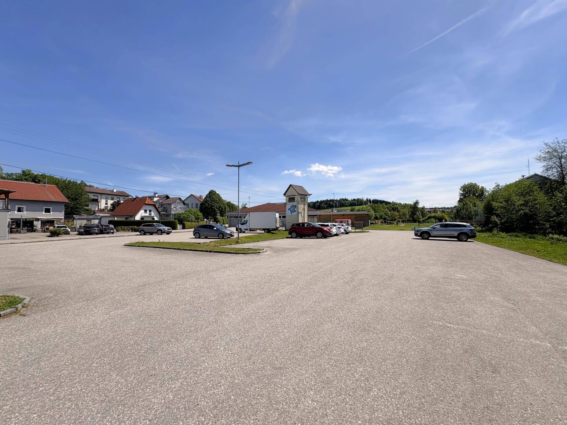 Großer asphaltierter Parkplatz mit umliegenden Gebäuden und Bäumen unter weitem blauem Himmel.