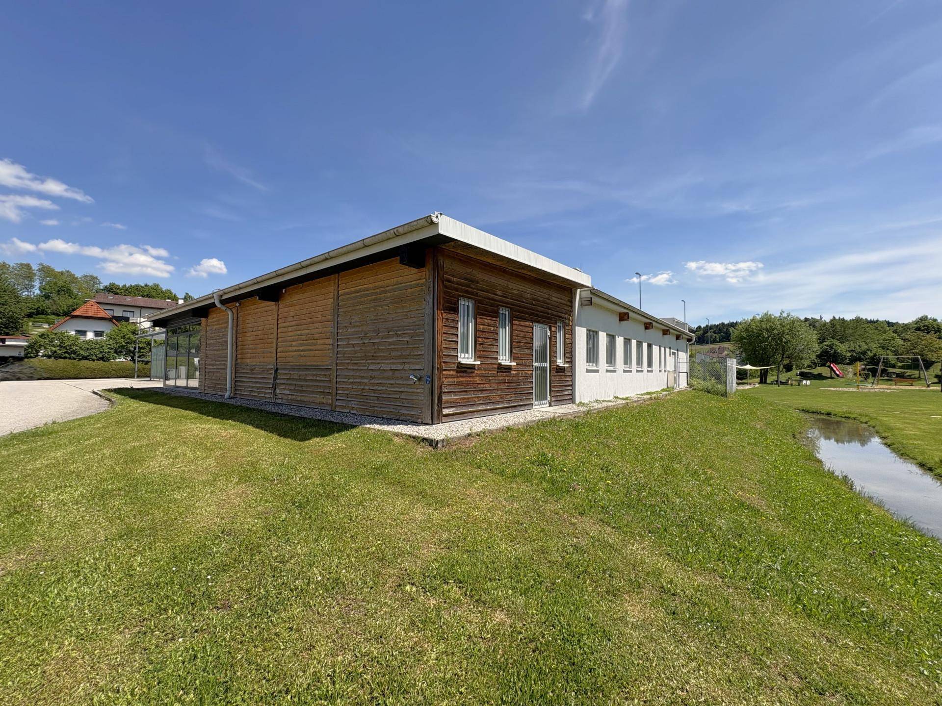 Seitenansicht des Gebäudes mit Holzverkleidung und weißer Fassade, angrenzend an eine Grünfläche mit Bach.