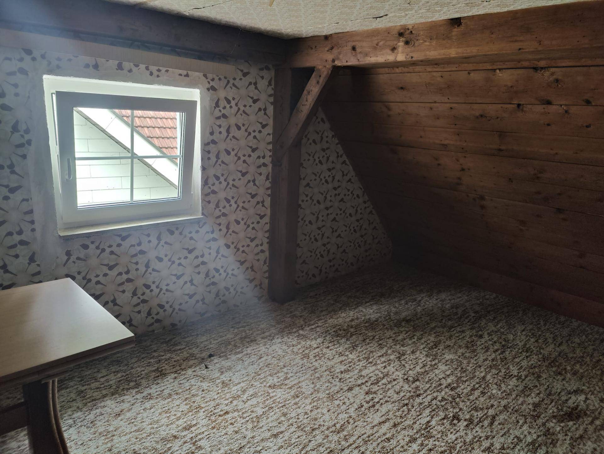 Dachzimmer mit Fenster, gemusterter Tapete, Teppichboden und sichtbaren Holzbalken, bietet gemütlichen Rückzugsort.