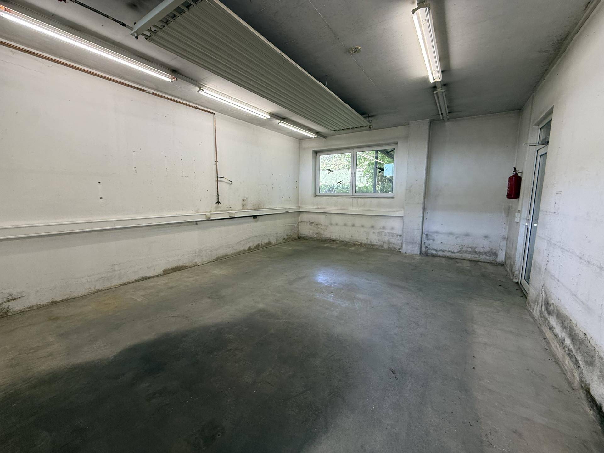 Kleinerer Lagerraum mit Betonboden, einem Fenster und sichtbaren Wasserleitungen an der Wand.