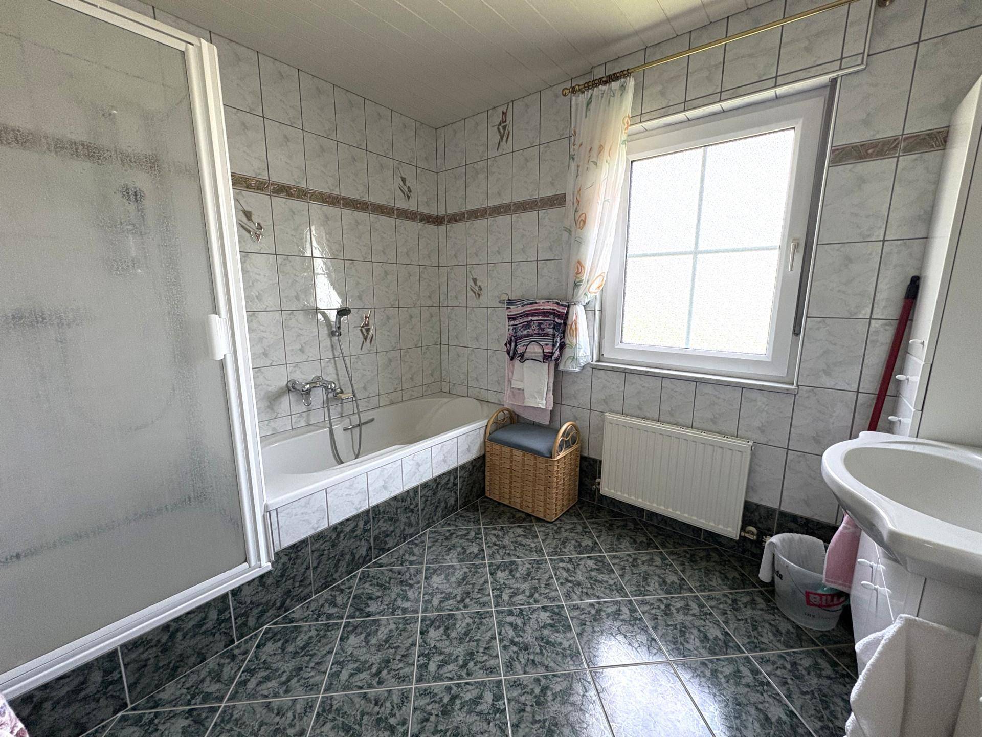 Badezimmer mit Badewanne, Duschkabine, Fenster und grauen Fliesen.