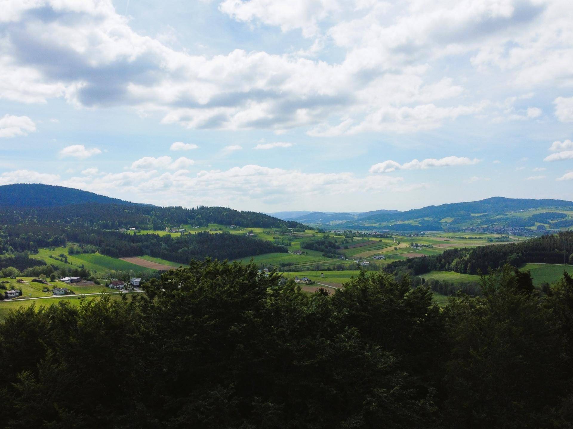Weitläufige Landschaft mit sanften grünen Hügeln, Feldern und kleinen Dörfern unter einem bewölkten Himmel.