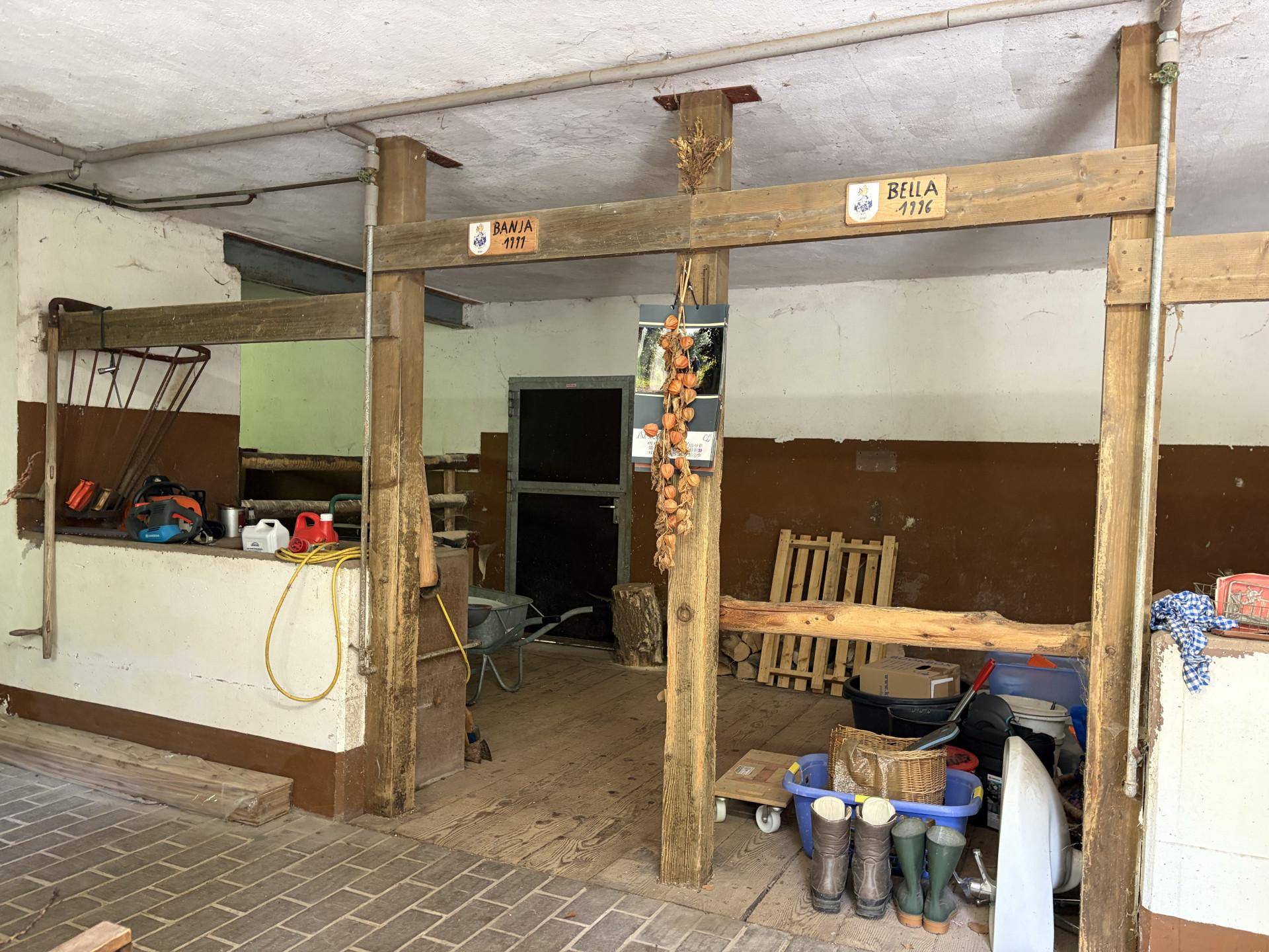 Rustikaler Innenraum mit Holzbalken und Betonboden, genutzt als Abstell- oder Stallbereich mit alten Beschriftungen.