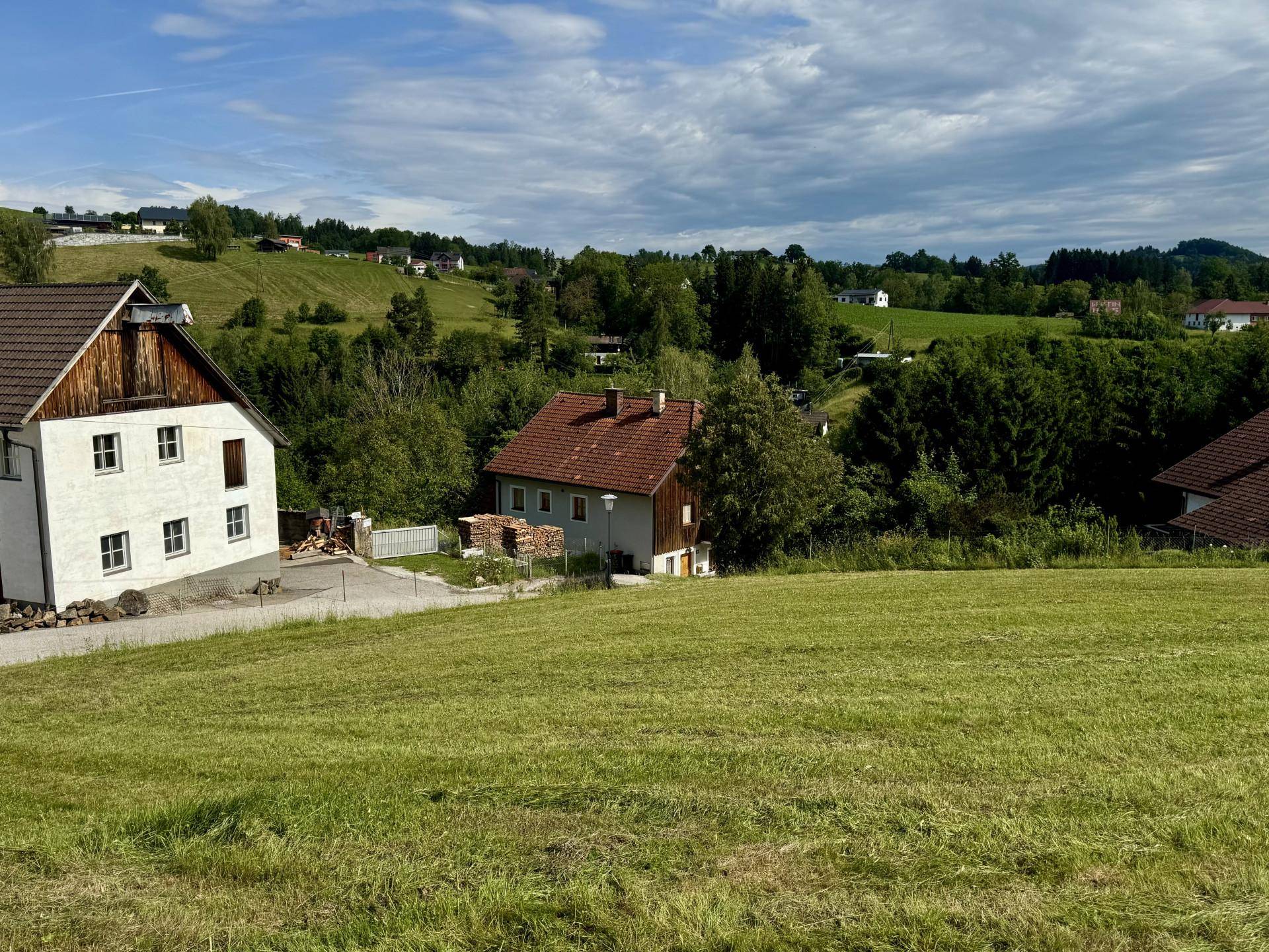 Panoramablick über eine weite grüne Wiese auf zwei Wohnhäuser und die idyllische Hügellandschaft.