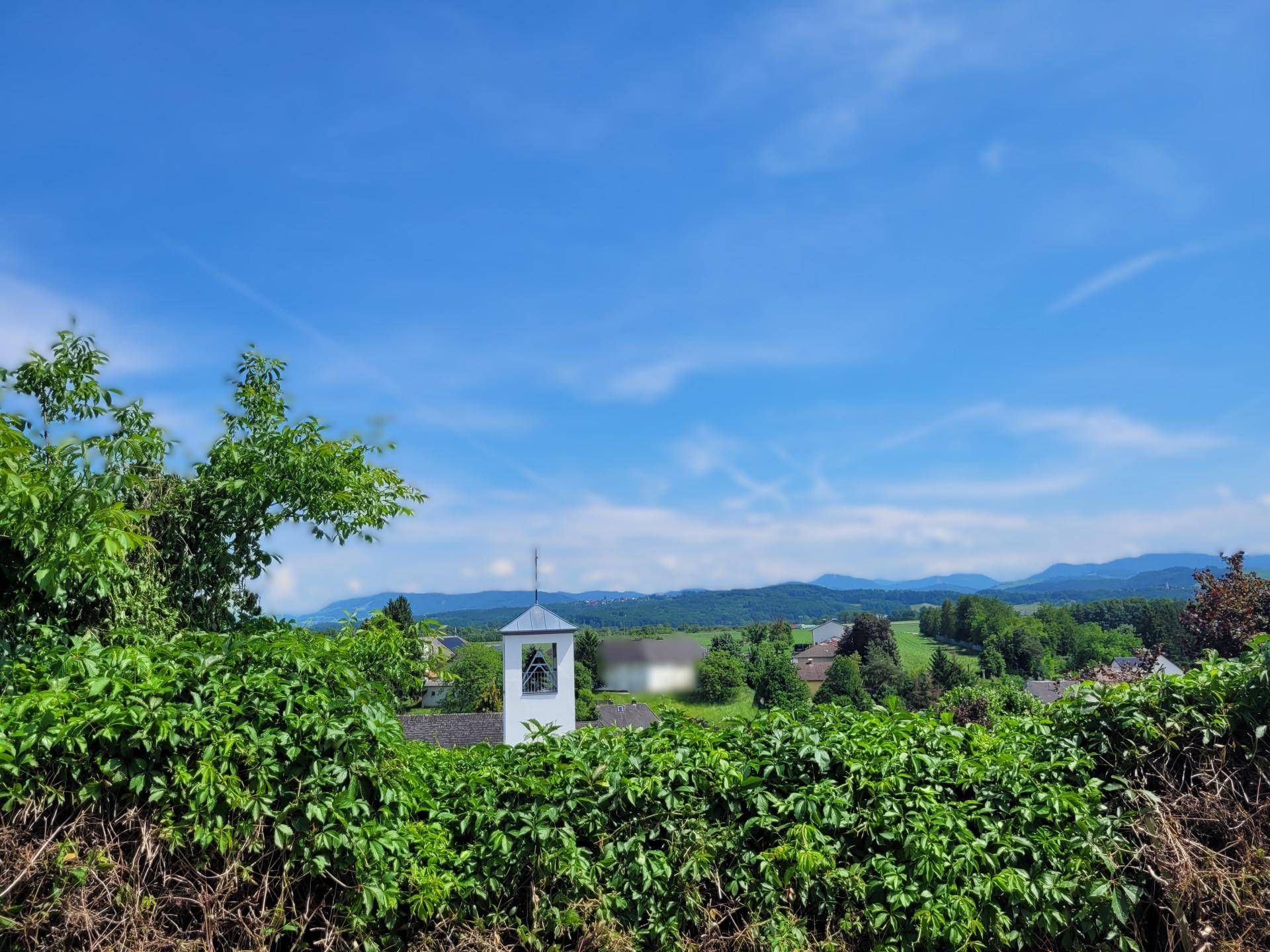 Weitblick über eine grüne Landschaft mit einem kleinen Glockenturm und fernen Bergen unter blauem Himmel.