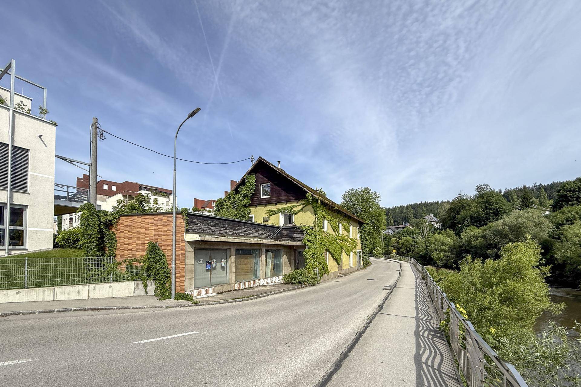 Straße mit einem alten, von Efeu bewachsenen Gebäude und einem modernen Bau, daneben ein Fluss.