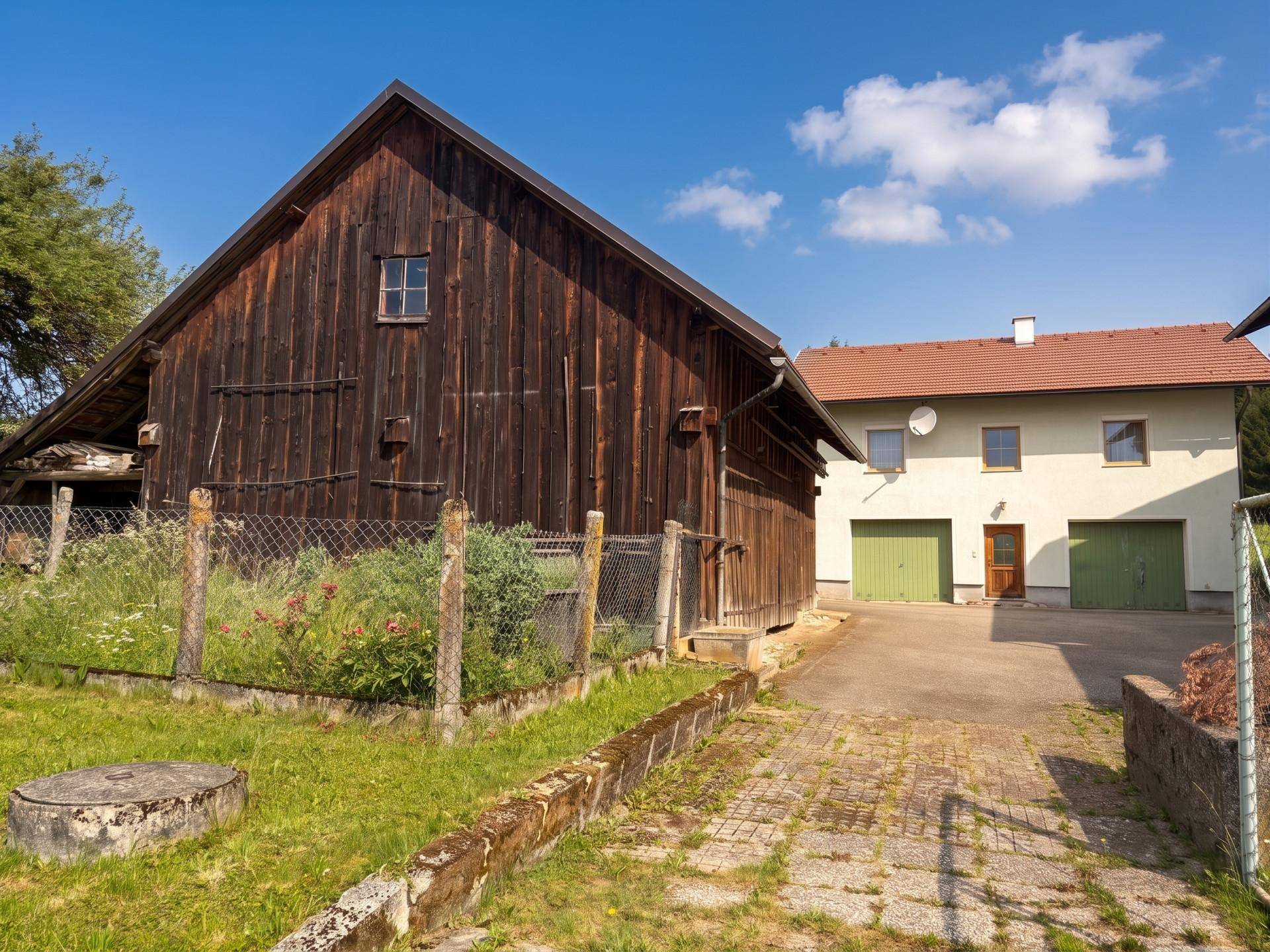 Rustikale Holzscheune neben einem Wohnhaus mit grünen Garagentoren und gepflasterter Zufahrt.