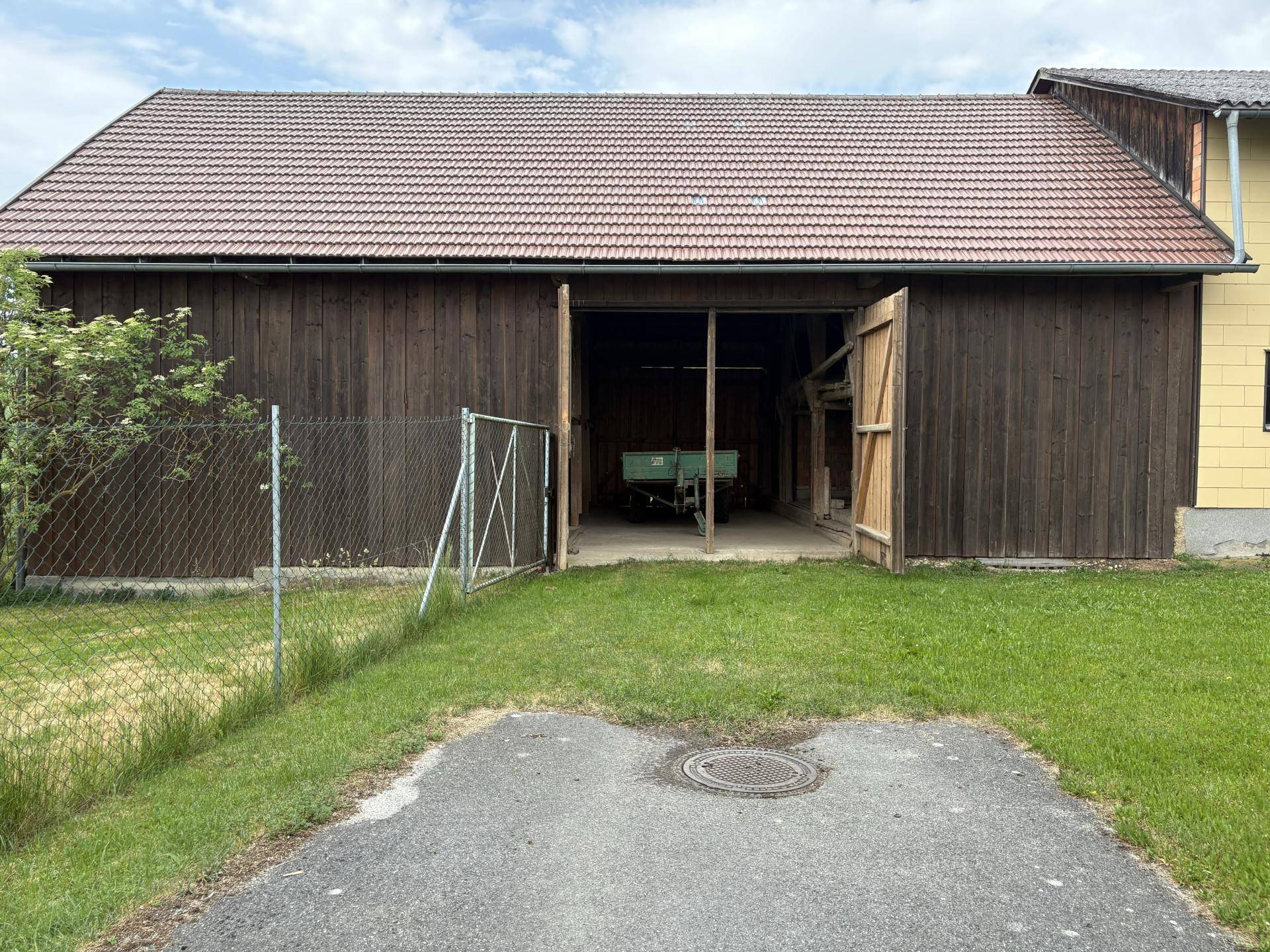 Große Holzscheune mit geöffneten Toren und Ziegeldach, umgeben von Gras und einem Zaun.