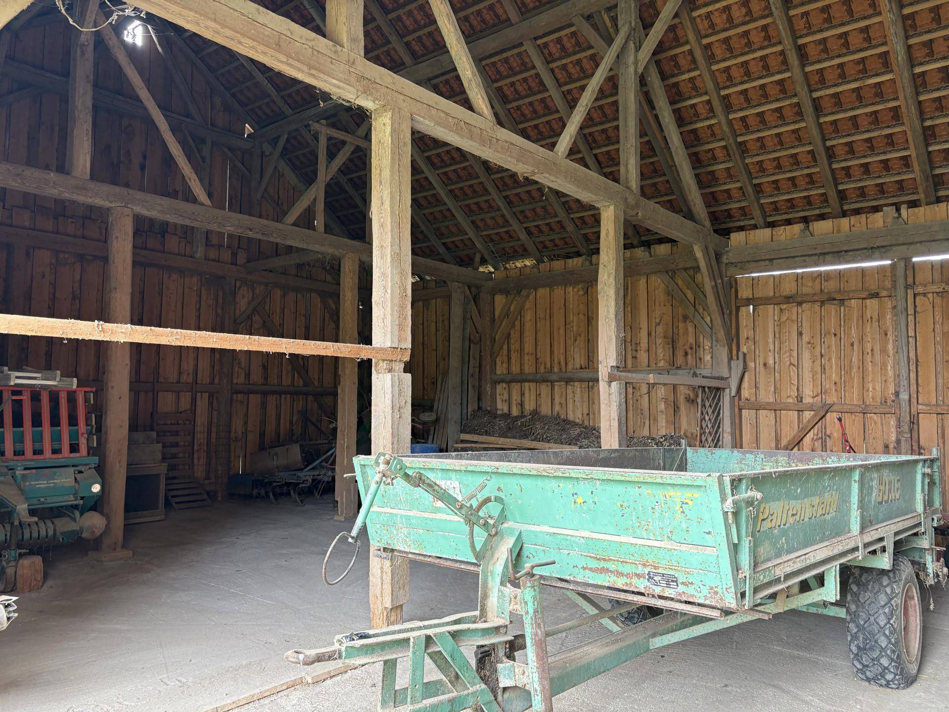 Geräumiger Innenraum einer Holzscheune mit sichtbaren Balken und einem landwirtschaftlichen Anhänger.
