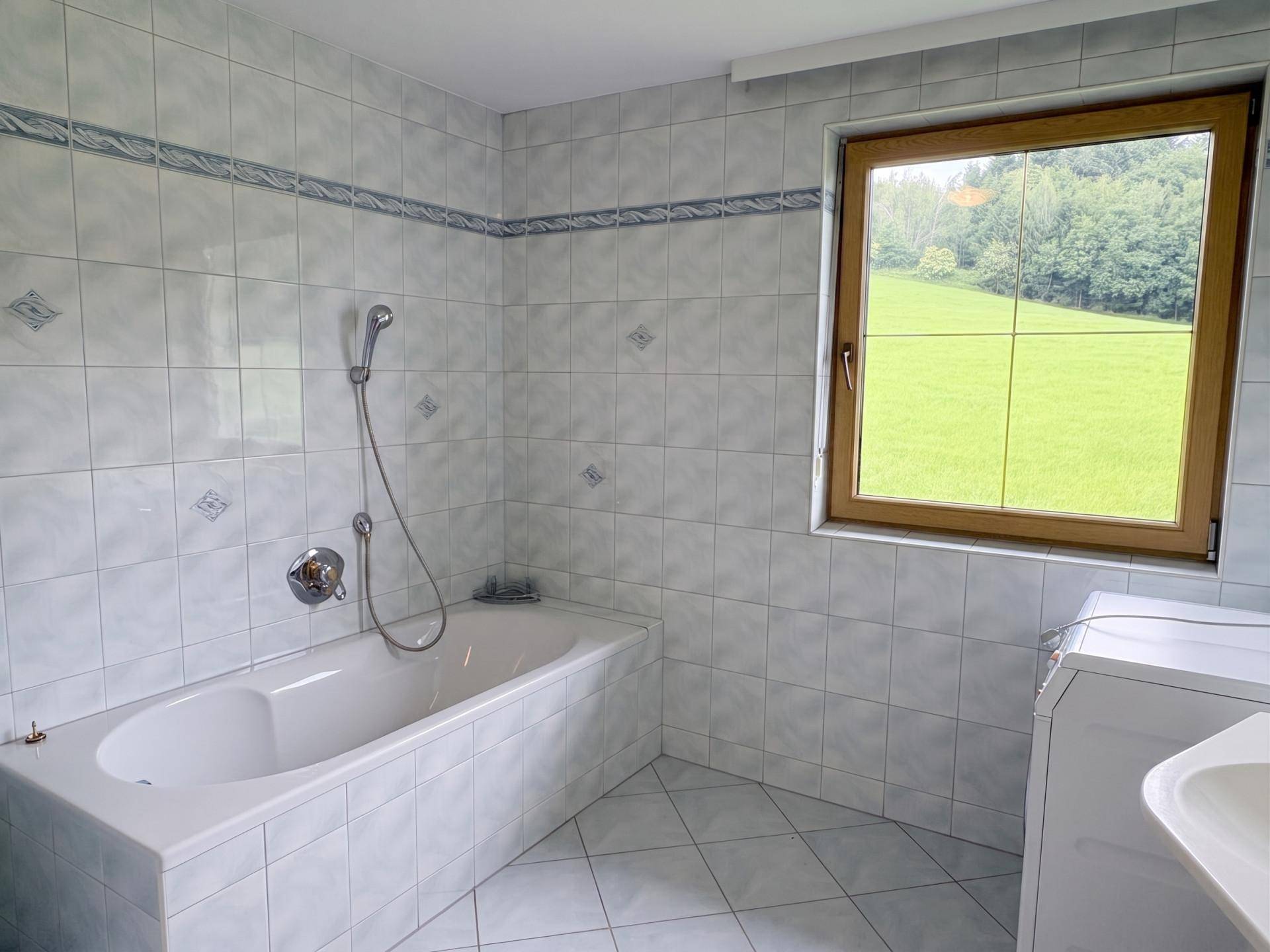 Gefliestes Badezimmer mit Badewanne, Waschbecken und einem Fenster mit Blick ins Grüne.