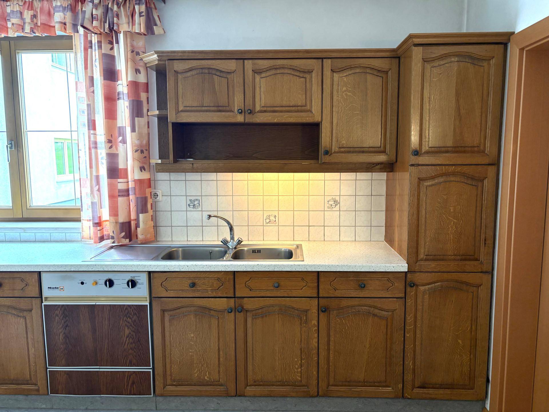 Traditionelle Holzküche mit Einbauschränken, Spüle und einem Fenster mit karierten Vorhängen.