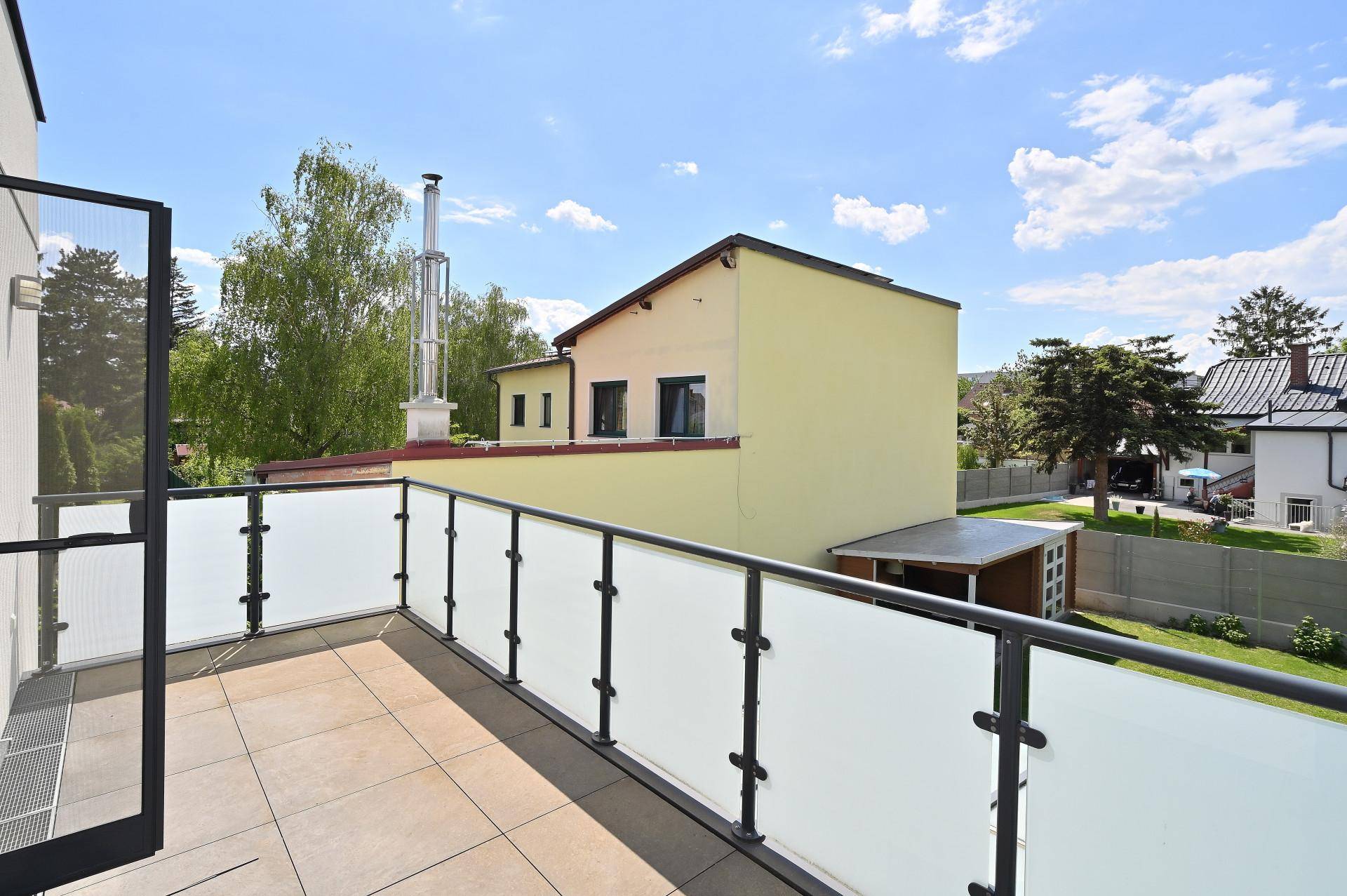 Sonniger Balkon mit Glasgeländer und Blick auf die Nachbarhäuser und den Garten.