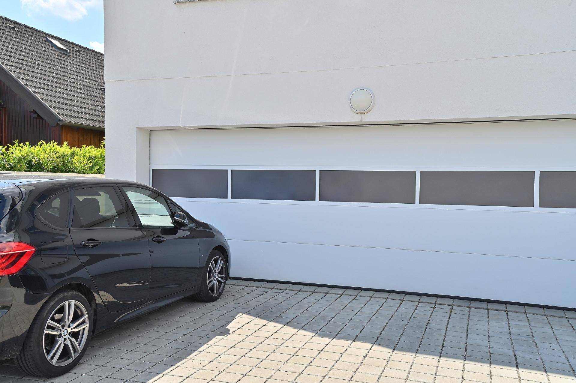 Moderne Garageneinfahrt mit einem schwarzen Auto davor und gepflastertem Vorplatz unter blauem Himmel.