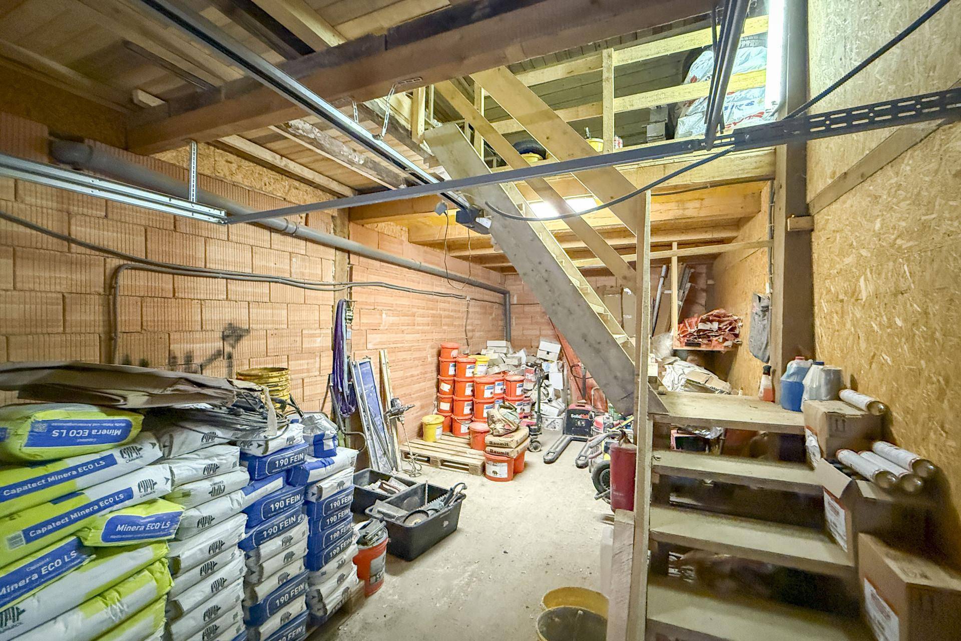 Lagerbereich mit Ziegelwänden, Säcken voller Baumaterialien und einer Holztreppe, die in ein Obergeschoss führt.