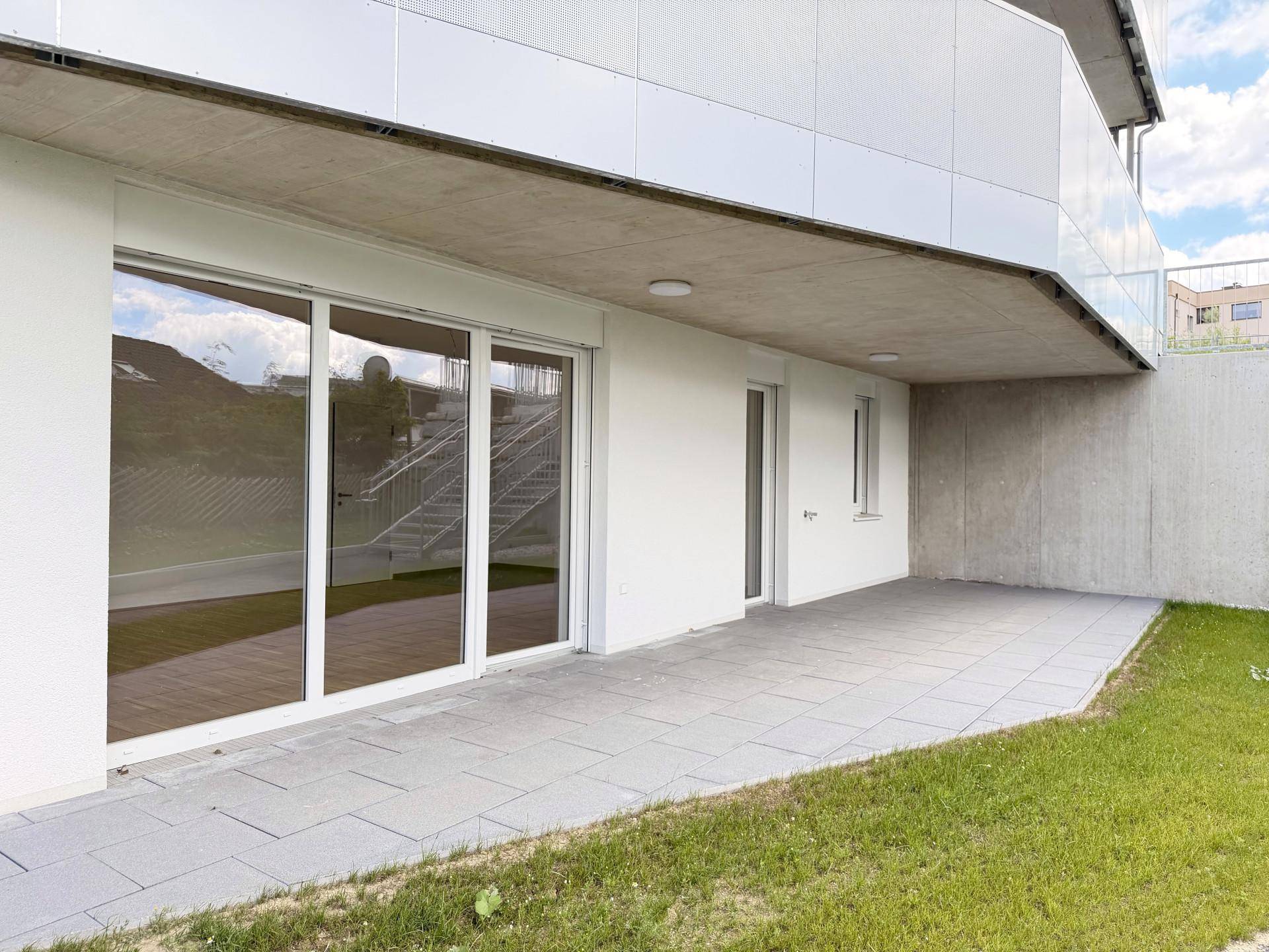 Großzügige Terrasse am Neubau mit Fliesenboden und angrenzender Rasenfläche, ideal für Entspannung.