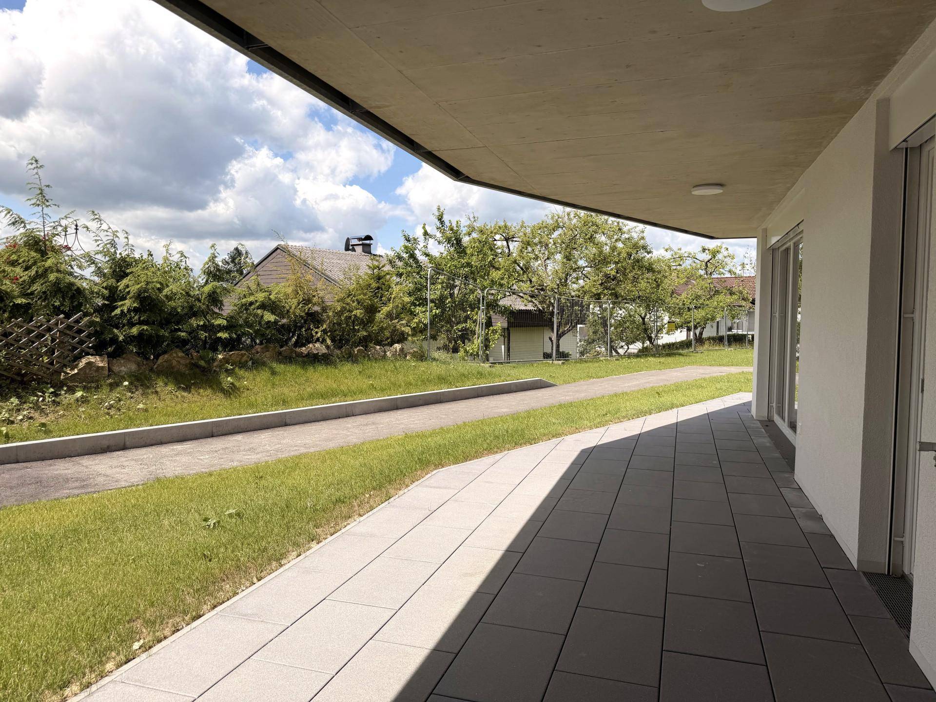 Überdachte Terrasse mit Fliesenboden und direktem Zugang zum Gartenbereich, umgeben von Grün.