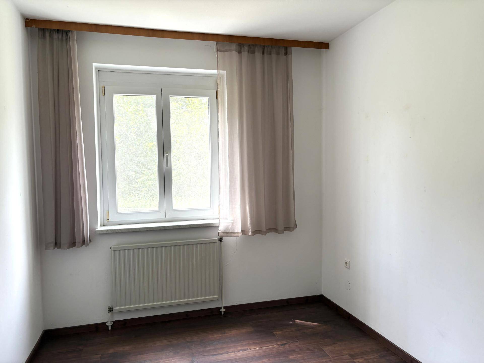 Leeres Zimmer mit Fenster, Heizkörper und dunklem Laminatboden.