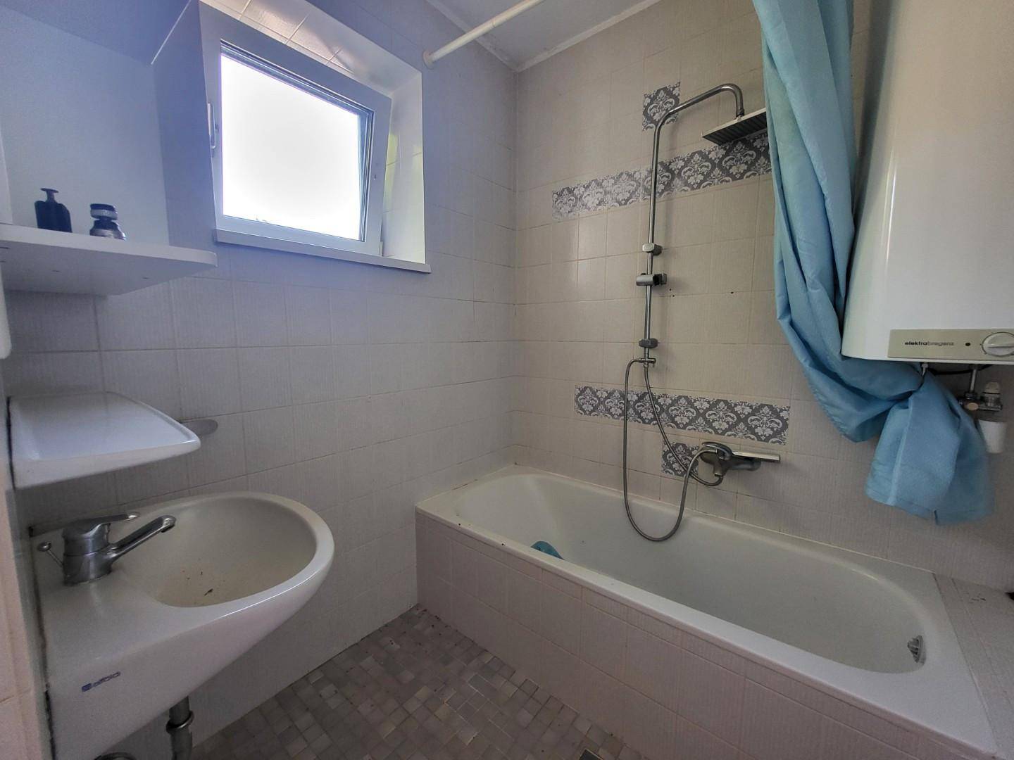 Badezimmer mit Badewanne, Dusche, Waschbecken und Fenster, in älterem Zustand.