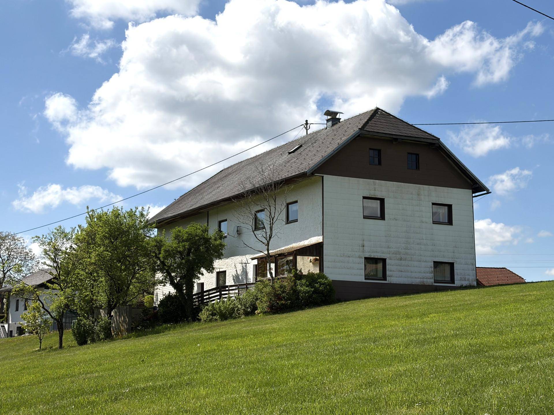Ländliches Wohnhaus mit weißer Fassade, braunen Fensterrahmen und dunklem Dach, eingebettet in grüne Landschaft.