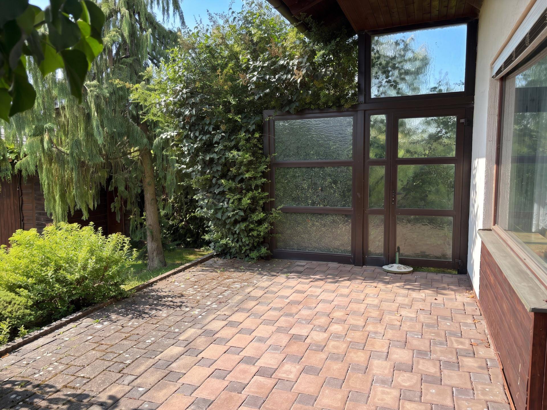 Gepflasterte Terrasse neben dem Haus, umgeben von Sträuchern und Bäumen, ideal für entspannte Stunden im Freien.