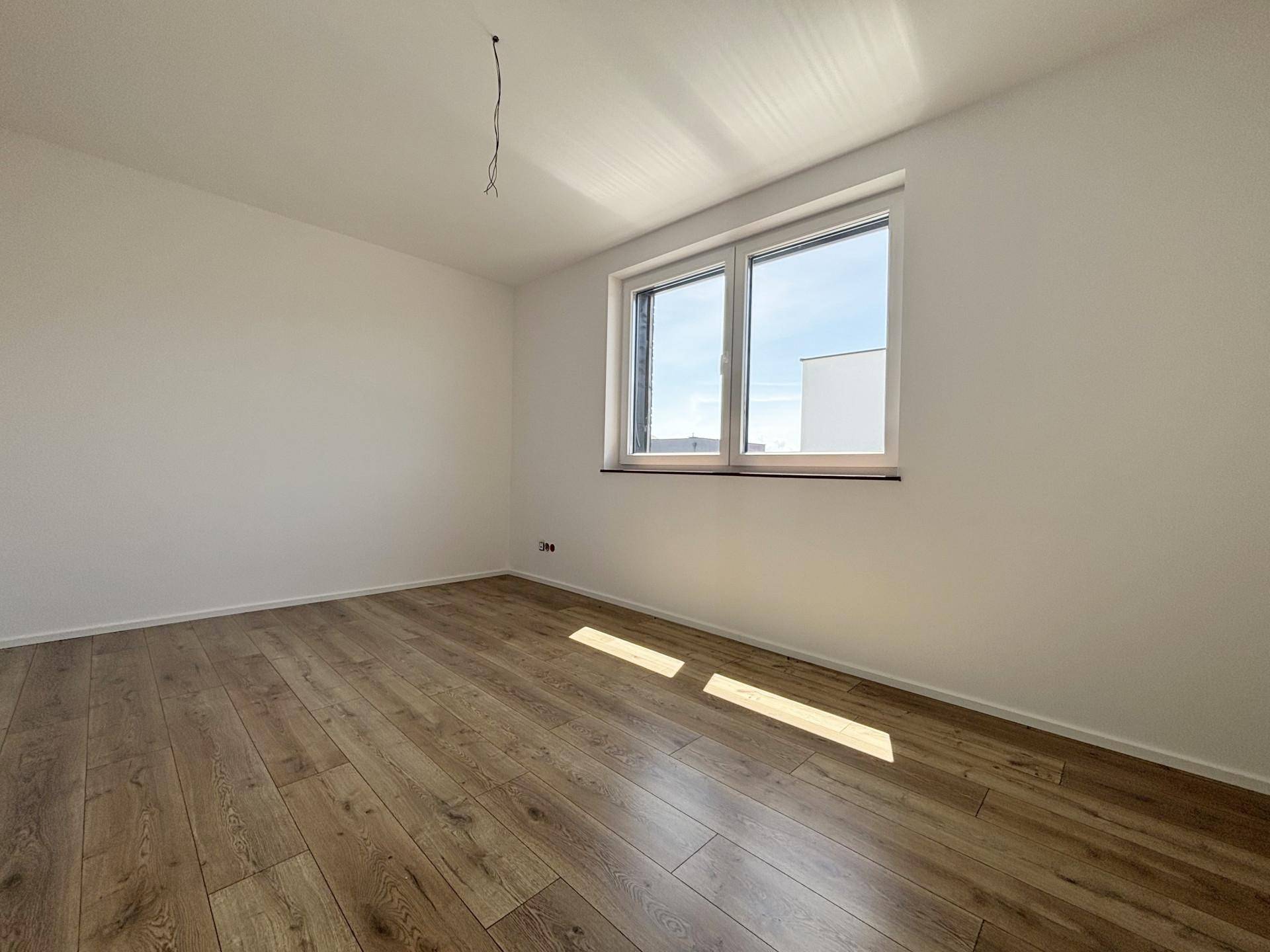 Geräumiges Zimmer mit Holzboden und großem Fenster, das viel Sonnenlicht hereinlässt.
