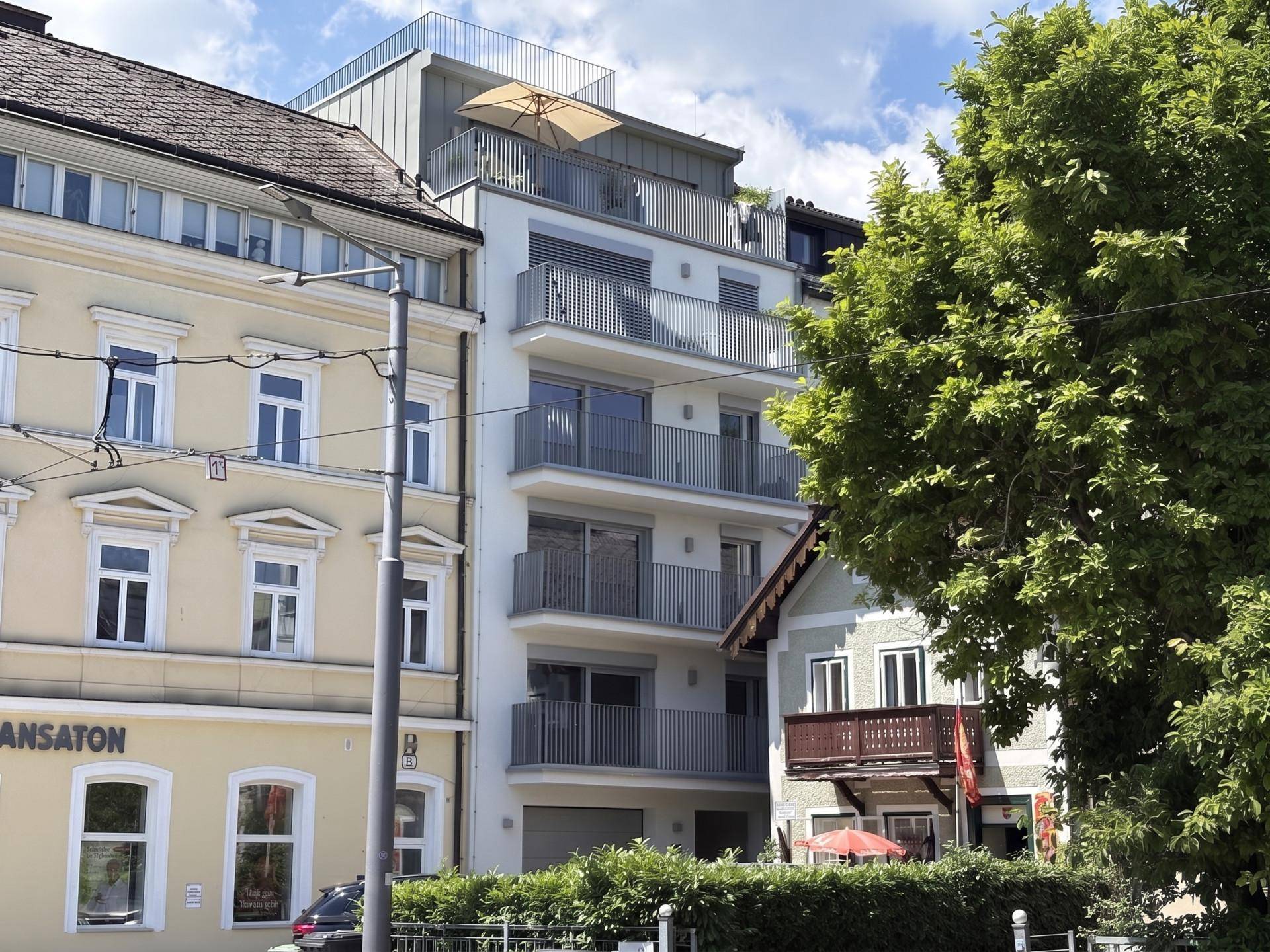 Modernes Mehrfamilienhaus mit Balkonen und Dachterrasse in städtischer Umgebung, umgeben von älteren Gebäuden.
