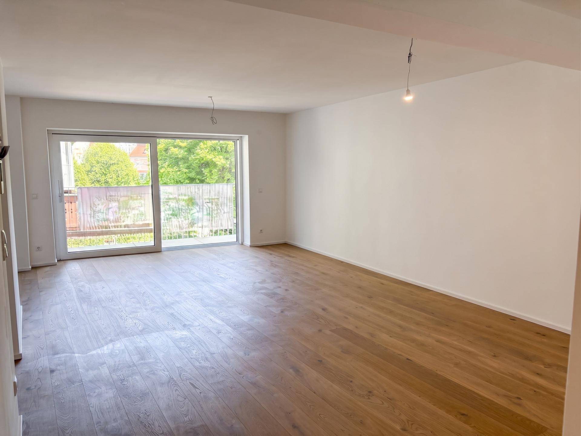 Heller, leerer Wohnraum mit Holzboden und großem Fenster mit Zugang zu einem Außenbereich.