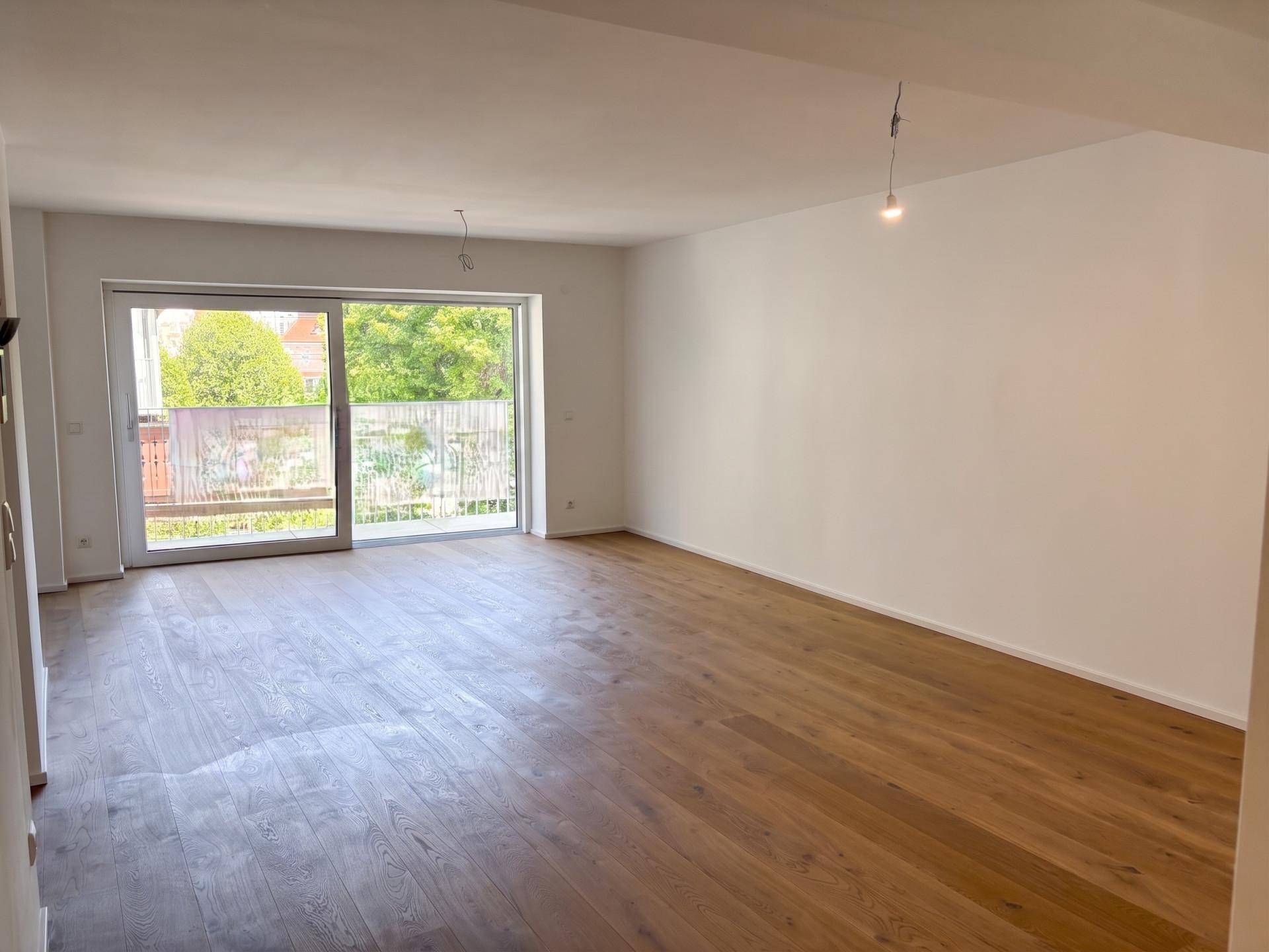 Geräumiger, unmöblierter Wohnbereich mit Parkettboden und Zugang zu einem Balkon mit Baumblick.
