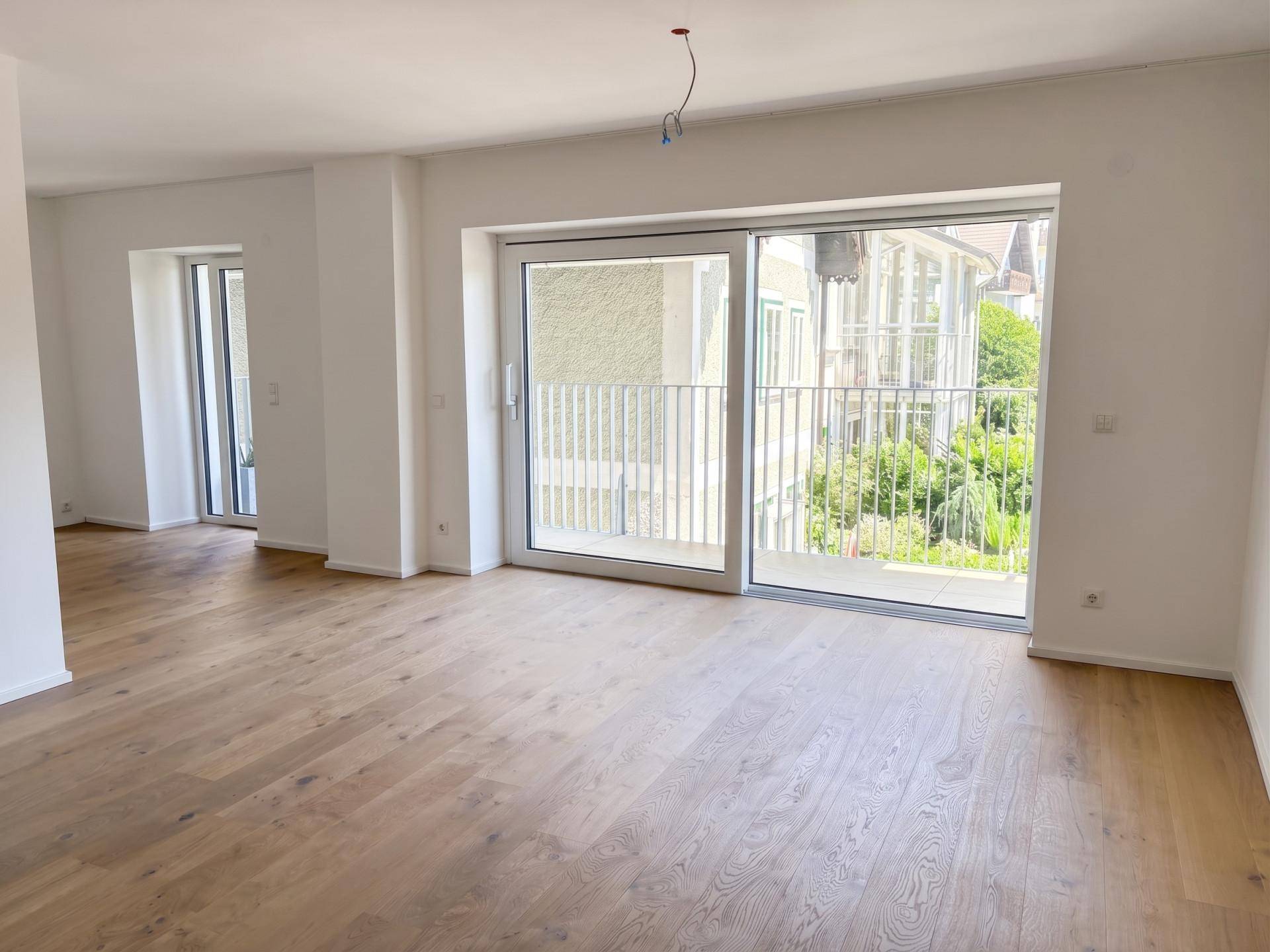 Heller, leerer Wohnraum mit Holzboden und großer Schiebetür zum Balkon mit Blick ins Grüne.