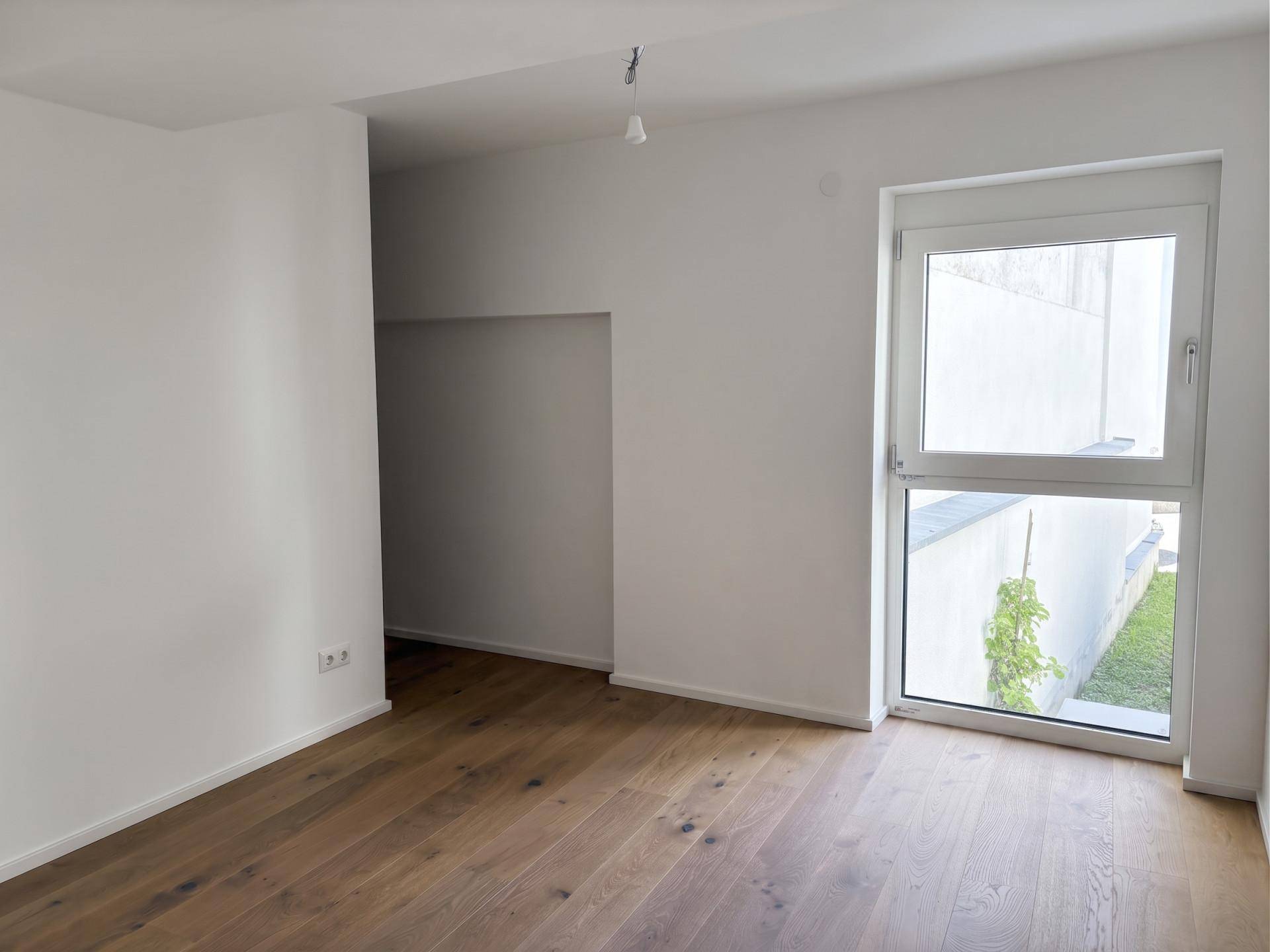 Heller, leerer Raum mit Holzboden und Fenster mit Blick auf einen kleinen Außenbereich.