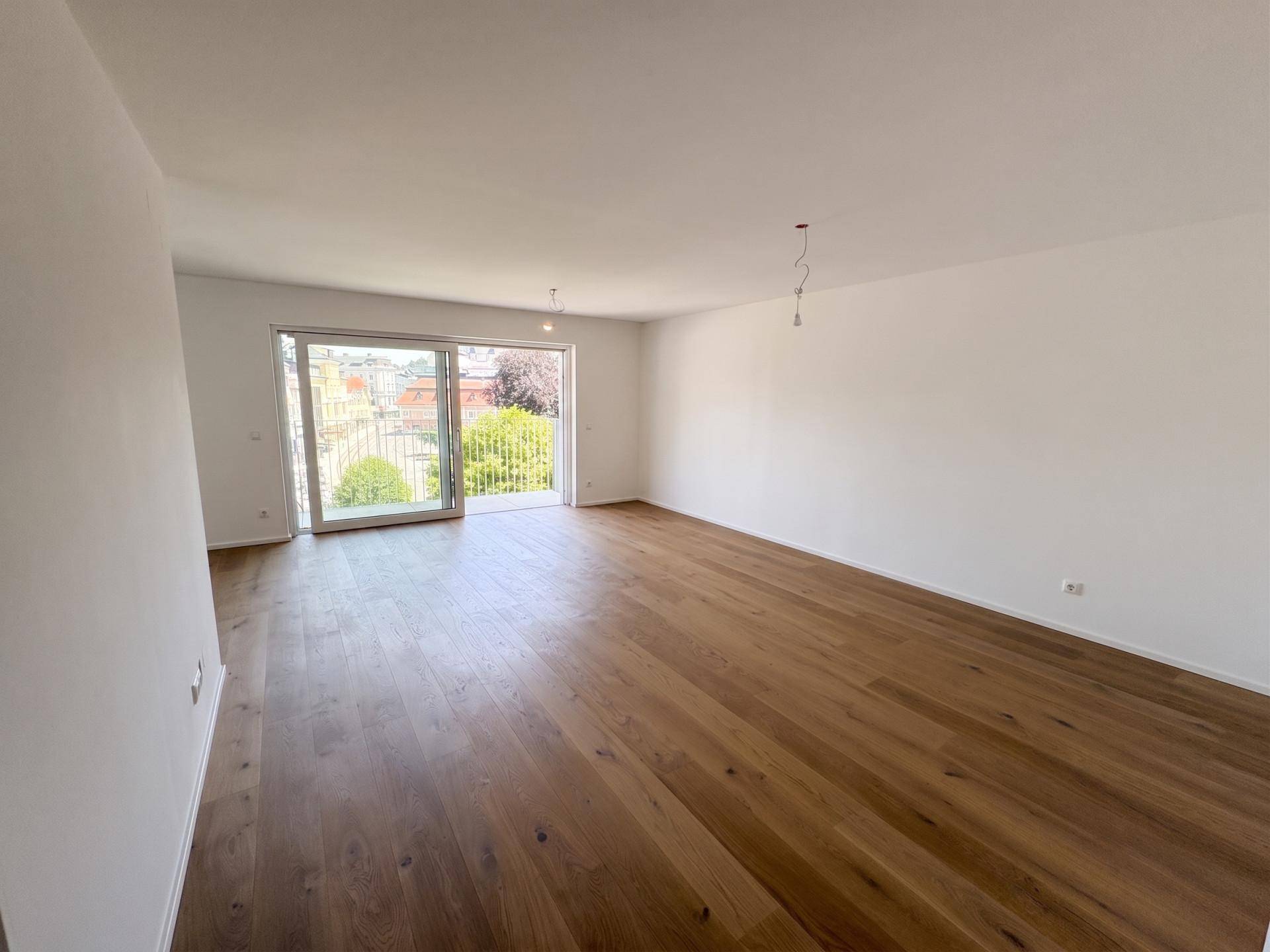 Geräumiger, unmöblierter Wohnbereich mit elegantem Holzboden und viel Tageslicht durch die Balkontür.