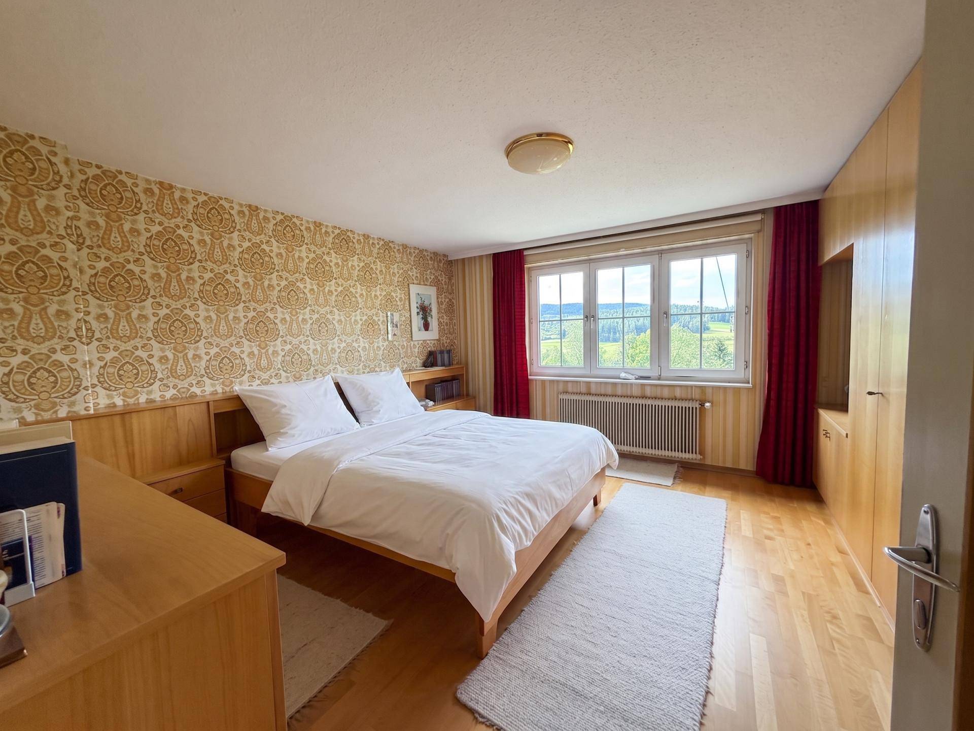 Schlafzimmer mit gemusterter Tapete, Holzboden und weitem Blick in die Landschaft.