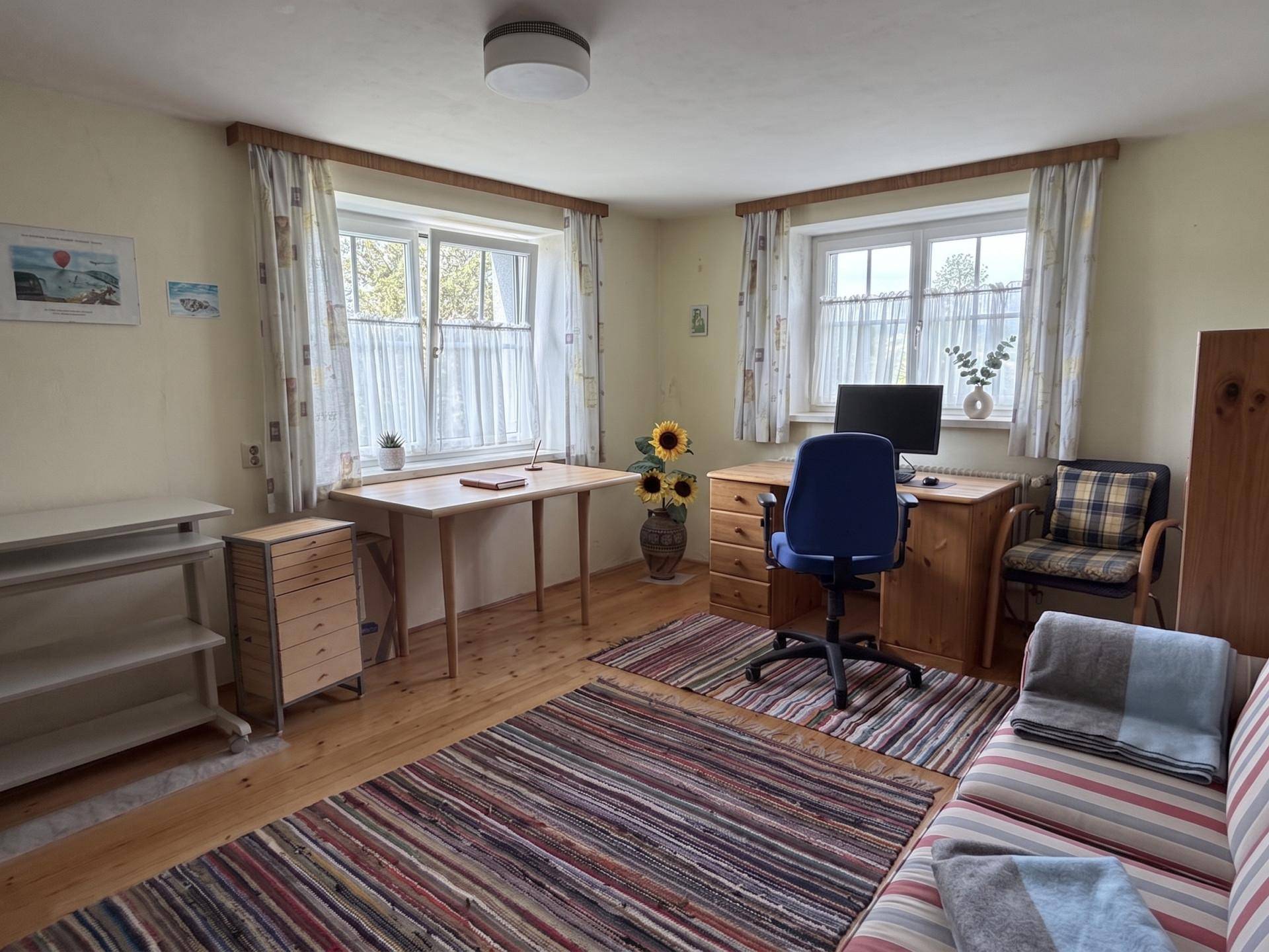Helles Arbeitszimmer mit Holzmöbeln, Teppich und zwei Fenstern für viel Tageslicht.