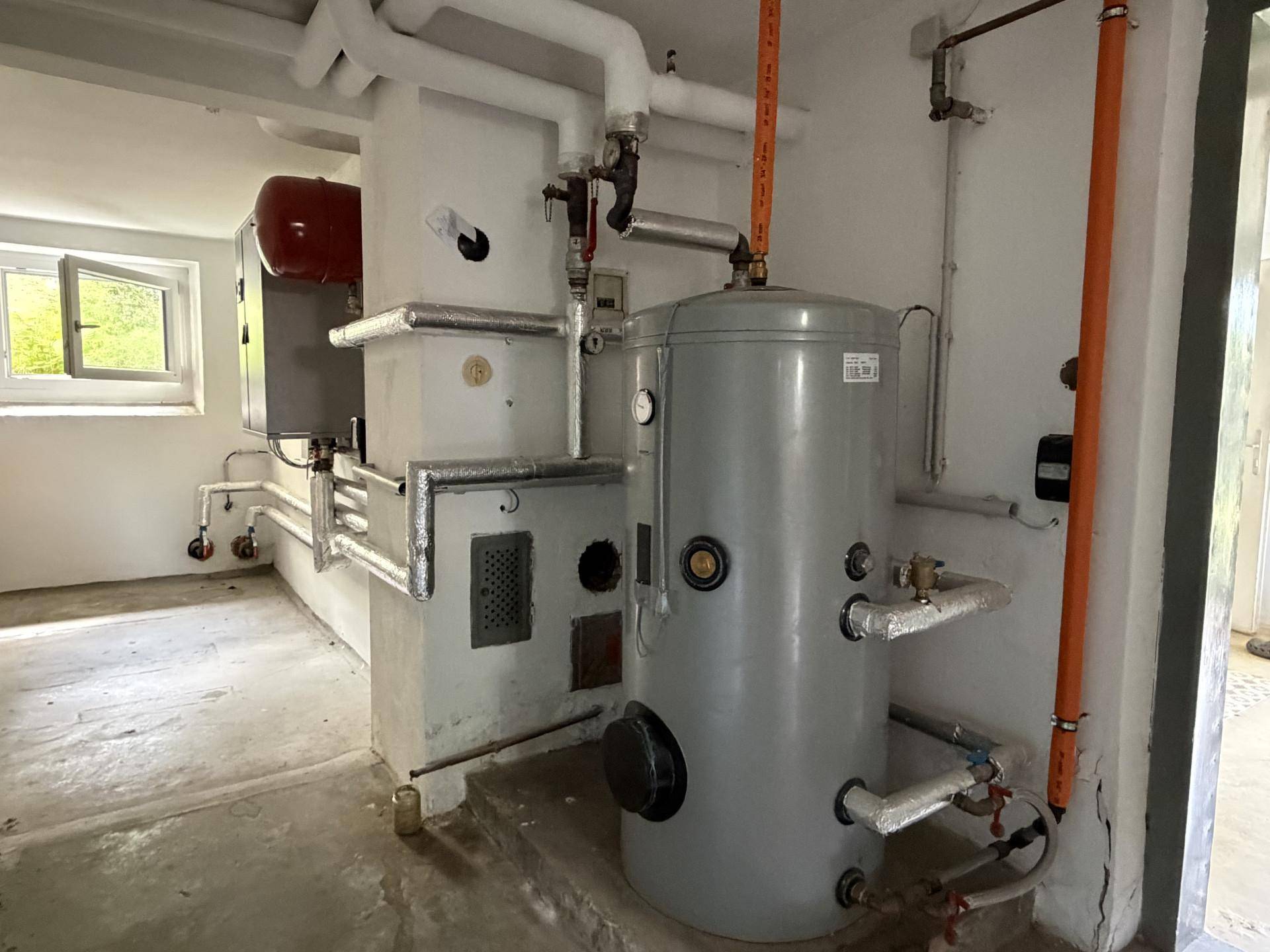 Heizungsraum im Keller mit Warmwasserspeicher und sichtbaren Rohrleitungen.