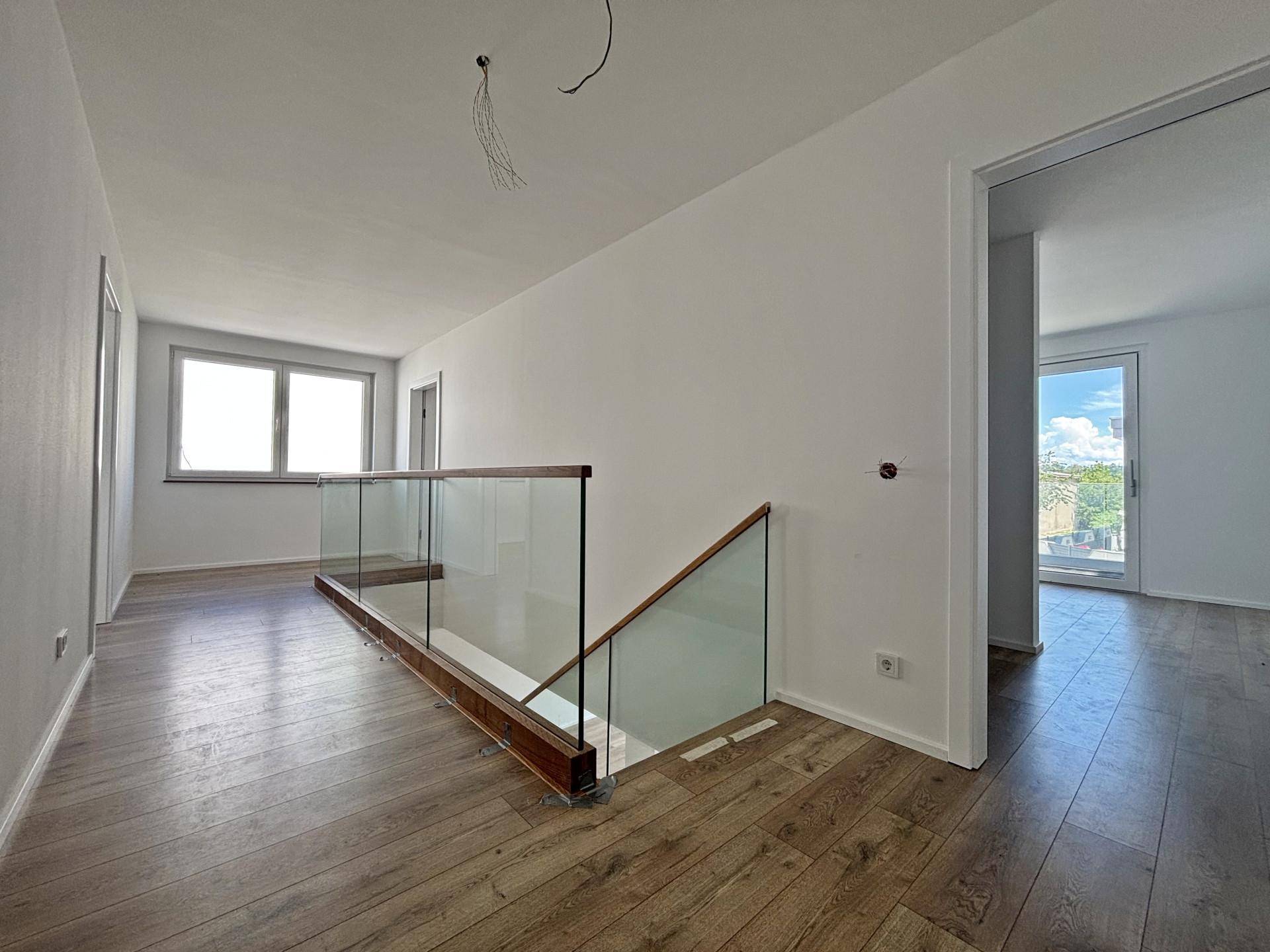 Heller Flur im Obergeschoss mit modernem Glasgeländer und Laminatboden, der zu weiteren Zimmern führt.