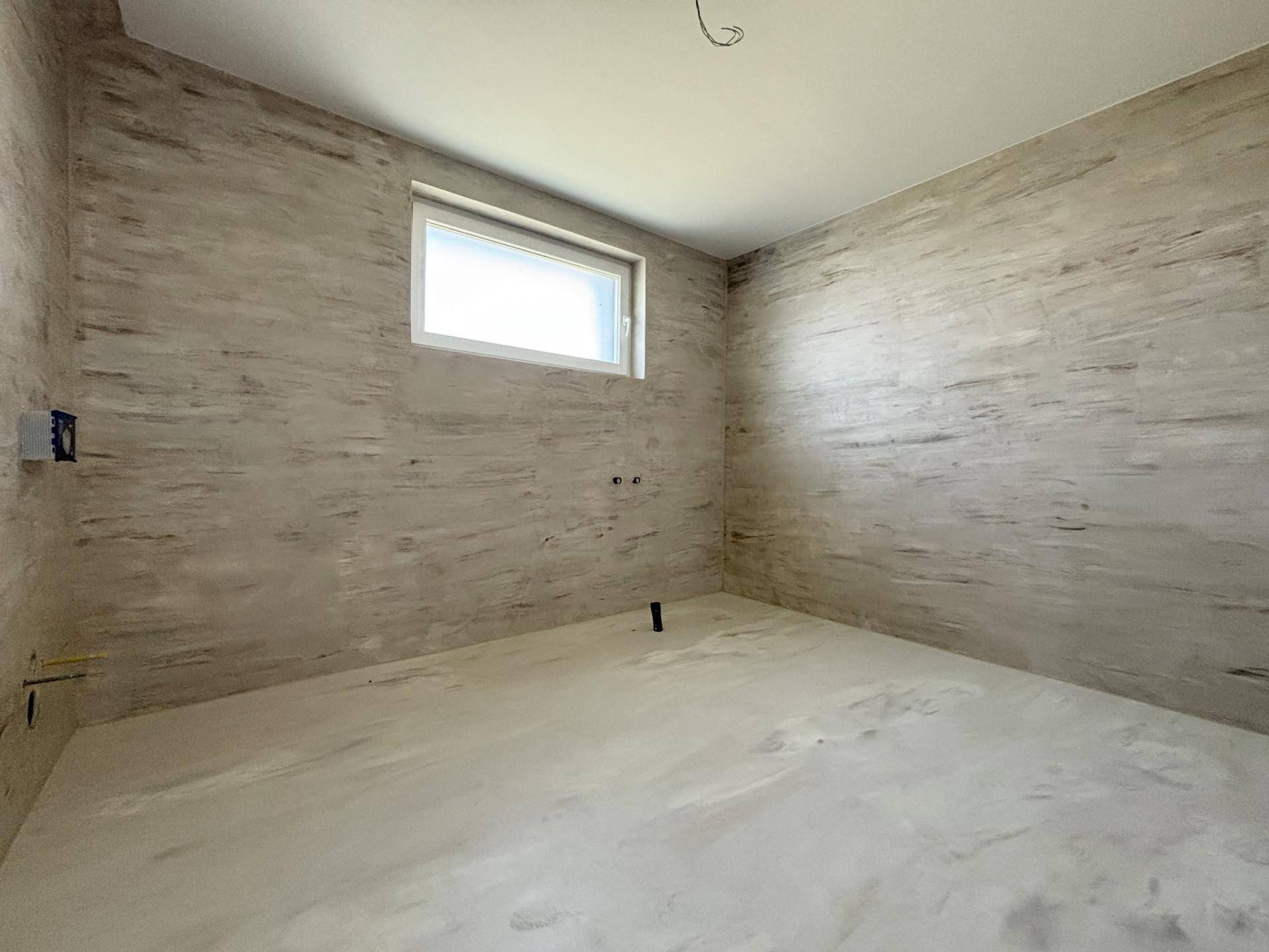 Unfertiges Badezimmer mit Fenster und vorbereiteten Anschlüssen für Sanitärinstallationen.