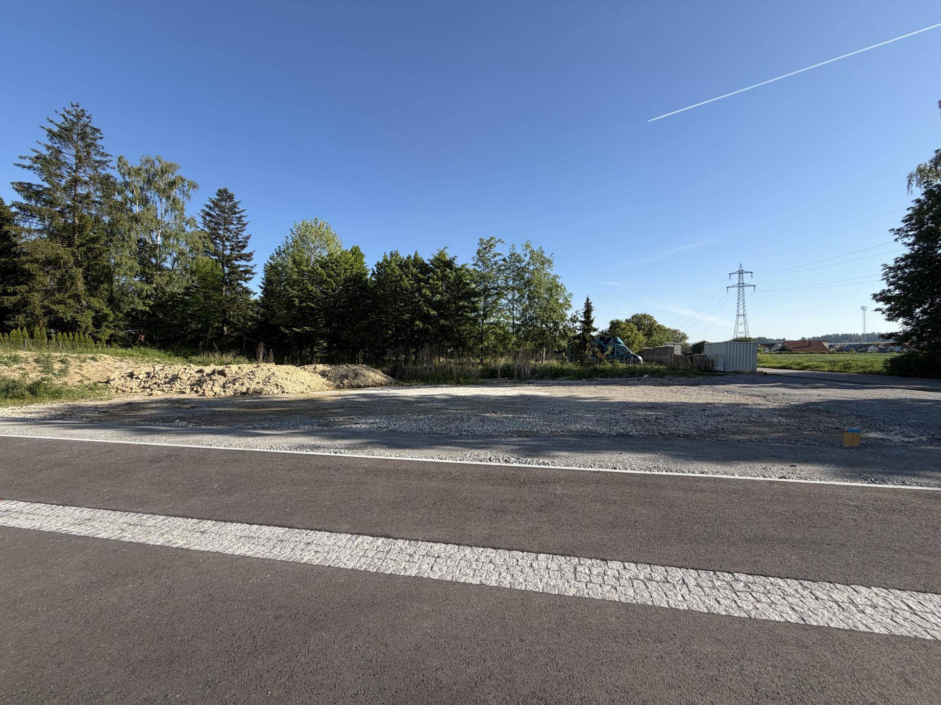 Unbebautes Grundstück mit Schotterfläche und Bäumen im Hintergrund unter klarem, sonnigem Himmel.